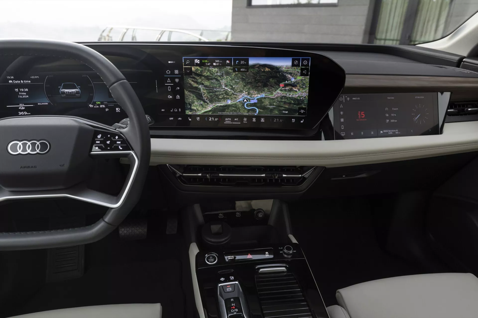 Interiören av en modern Audi Q6 e-tron 2024, sett från en förarperspektiv. Funktioner inkluderar en läderklädd tre-ekrad ratt med inbyggda kontroller, en digital instrumentpanel som visar hastighet och navigering, och ett horisontellt orienterat infotainmentsystem med pekskärm. Mittkonsolen inkluderar kontroller för klimatinställningar och körlägen. Lyxiga material som läder och träpaneler förhöjer estetiken.