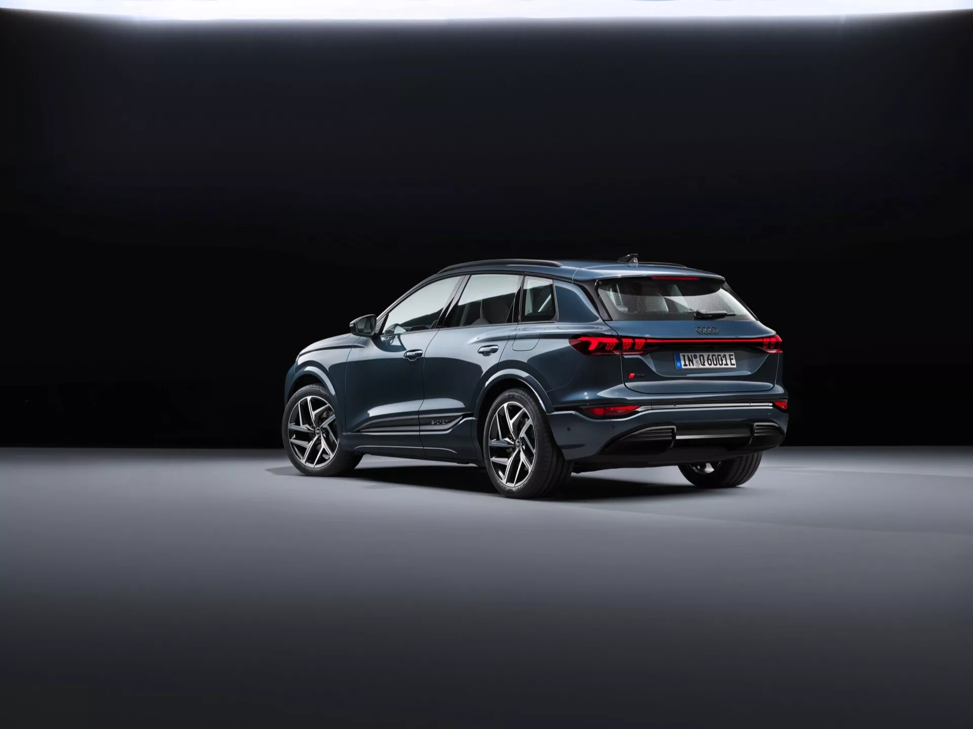 Den bakre trekvarts vyn av Audi Q6 e-tron 2024 (gf) visar en modern SUV placerad diagonalt på en slät, reflekterande showroom-golv. Bakgrunden övergår från mörkt till ljust, vilket framhäver fordonet. Synliga funktioner inkluderar sammankopplad bakre ljusenhet, särskiljande takspoiler, uttalade hjulhus med stora lättmetallfälgar, kromlist längs fönsterkanterna, en bakre stötfångare med reflektorer och sensorer samt ett dubbelt avgasrörssystem som antyder prestanda, vilket indikerar att det kan vara en hybrid- eller elmodell.