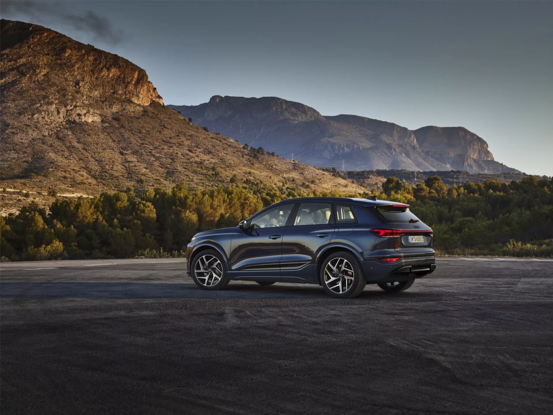 Bilden visar en modern Audi Q6 e-Tron 2024 SUV i en utomhusmiljö med pittoresk bergsbakgrund under gryning eller skymning. Den trekvartsvinklade bakifrån framhäver de distinkta LED-bakljusen, den rena bakluckedesignen som möjligen har en backkamera och ett dubbelt avgasrör. Sidprofilen visar upp de flerspåkiga aluminiumfälgarna, stora fönster och en lutande taklinje, vilket antyder en sport- eller lyxmodell med möjliga aerodynamiska funktioner. Sidospeglarna kan potentiellt integrera döda vinkeln-övervakningsteknologi. Det naturliga ljuset förstärker fordonets äventyrliga och kapabla attraktionskraft.
