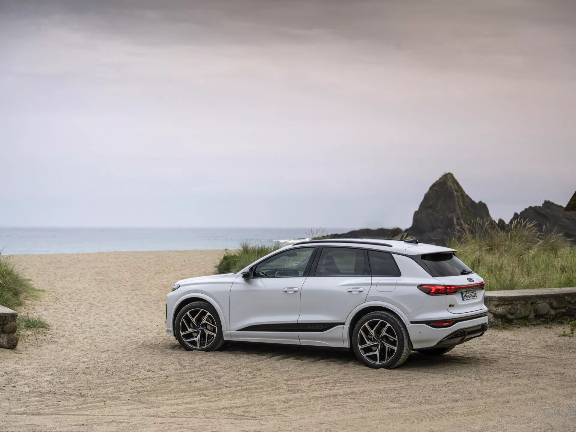 En vit Audi Q6 e-tron 2024 SUV är parkerad på en grusväg med en strandliknande bakgrund, fångad från en bakre trekvartsvy. Bilden framhäver fordonets moderna designelement inklusive baklyktor, bakre stötfångare, stiliga hjul och eleganta linjer. Bilens högre markfrigång och allmänna hållning indikerar dess lämplighet för olika terränger. Mjuk, jämn belysning antyder en mulen dag eller nära soluppgång/solnedgång, vilket förstärker den äventyrliga omgivningen. Moderna lättmetallfälgar, en bakre spoiler och LED-baklyktor är synliga, vilket signalerar avancerad teknik och en sportig karaktär.