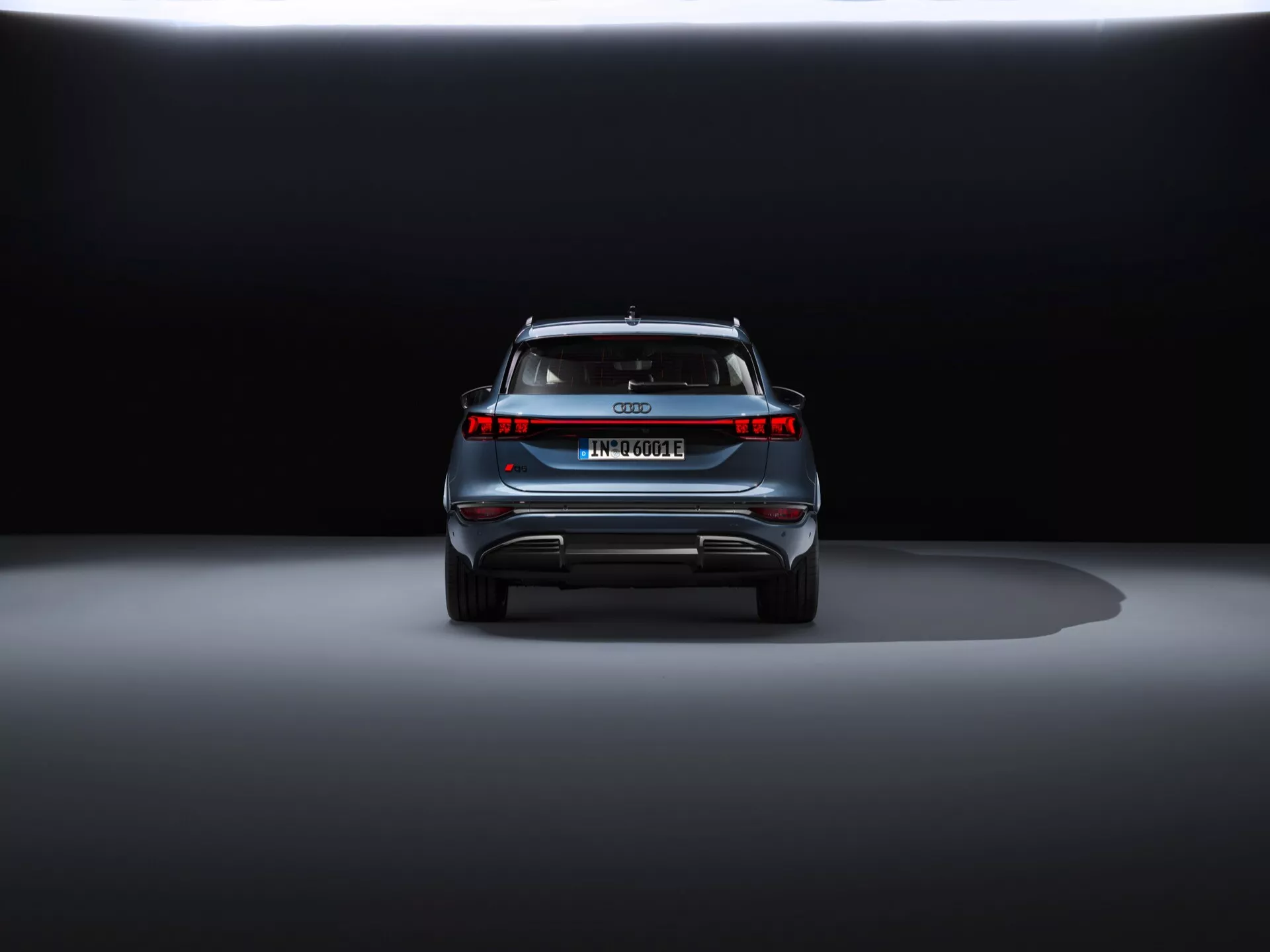 Bakre vy av Audi Q6 e-tron 2024 på utställning med dramatisk belysning. Marknivåbild framhäver moderna LED-baklys, avsaknad av avgasrör, fullbreddsljuslist, europeisk registreringsskylt, takspoiler och reflekterande säkerhetselement, vilket indikerar att det är ett lyxigt elektriskt eller hybridfordon
