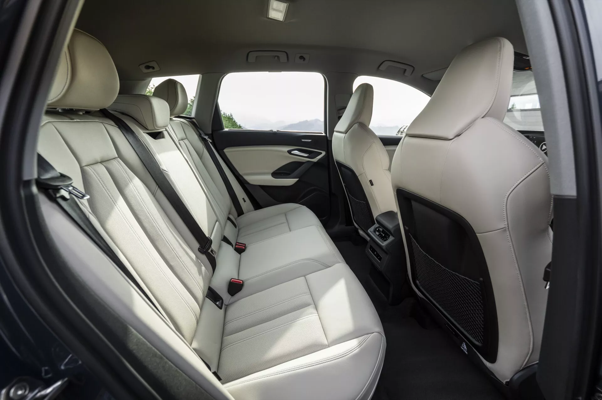 2024 Audi Q6 e-tron bakre passagerarkonruminteriör med fokus på baksätesarrangemang, ljusa lädersoffor med nackstöd, armstöd, säkerhetsbälten och spännen. Dörrpanel med högtalargaller, handtag och eventuellt fönsterkontroller. Färgtema i två toner med ljus och mörk trim. Luftkonditioneringsventiler mellan framsätena indikerar klimatkontroll för baksätena. Modern, lyxig och bekväm design för passagerare.
