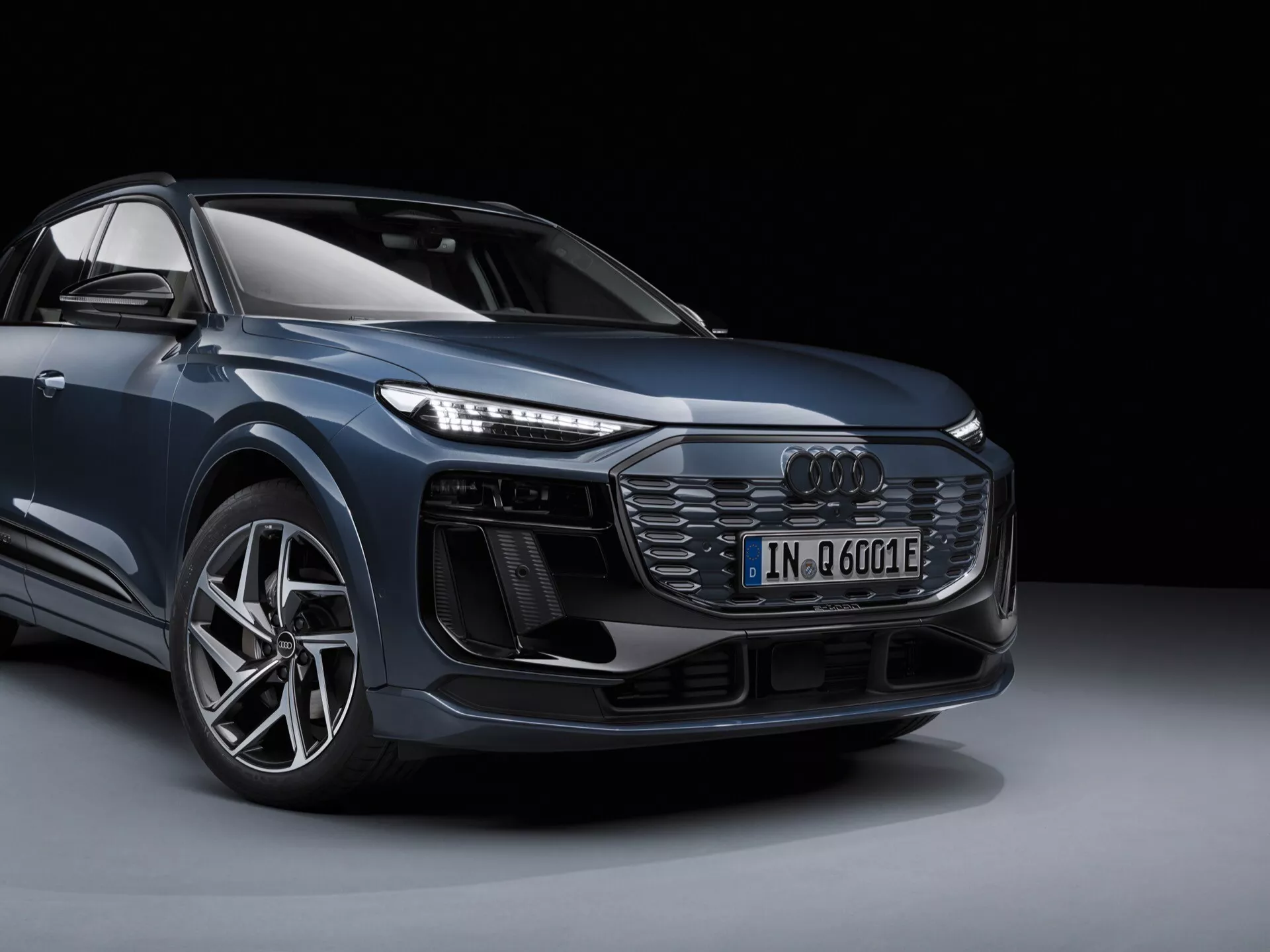Bilden visar en modern Audi Q6 e-tron 2024 i en studiomiljö, presenterad från en frontriktig trekvarts vy som erbjuder ett tydligt intryck av fordonets design framifrån och från sidan. Bilens vänstra främre hörn är framträdande, inklusive främre grillen med honungsrutemönster och centralt placerad Audi-logotyp, intrikata LED-framlyktor, ytterligare luftintag eller sensorer under framlyktorna, en sportig främre stötfångare, en stor lättmetallfälg med lågprofil däck samt konturen av en sidobackspegel. Bilden betonar den elektrifierade modellens emblem ('e') och framhäver dess design och tekniska egenskaper. Bakgrunden är enkel och mörk vilket säkerställer att inget avleder uppmärksamheten från fordonet självt.
