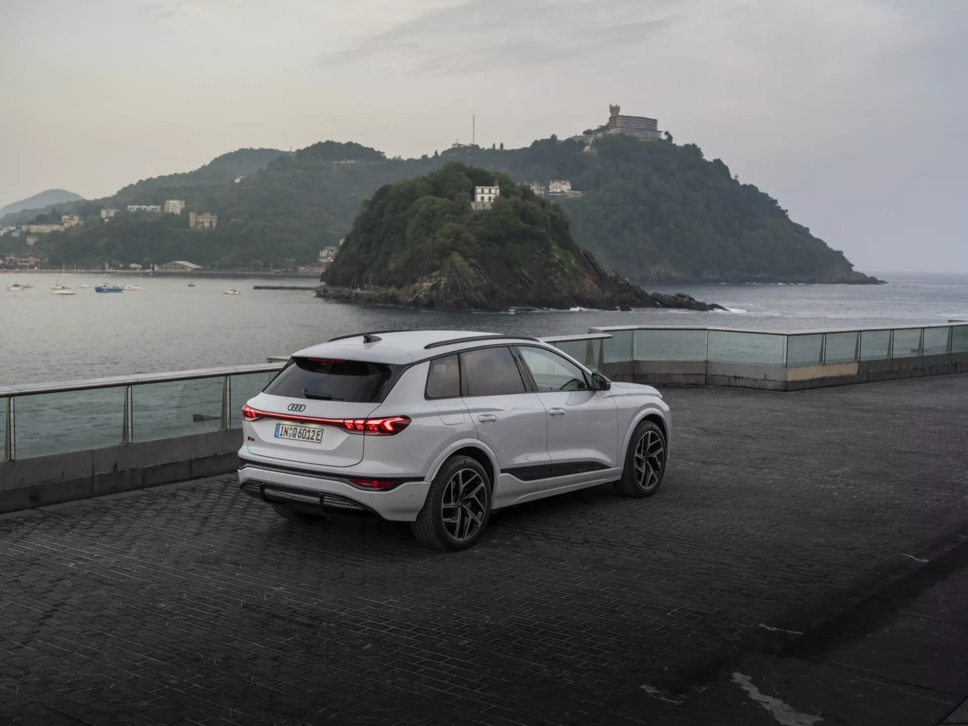 Trekvart bakifrån vy av en modern, vit Audi Q6 e-tron 2024 SUV parkerad på en sjöpromenad i skymningen, som visar sina horisontella bakre LED-ljus, tillverkarens emblem, bakre vindrutetorkare, femekrade lättmetallfälgar, integrerade blinkljus i sidospeglarna, takräcken och skulpturala karosslinjer mot en lugn bakgrund med en liten ö och en byggnad, vilket framhäver en blandning av funktionalitet och prestanda.
