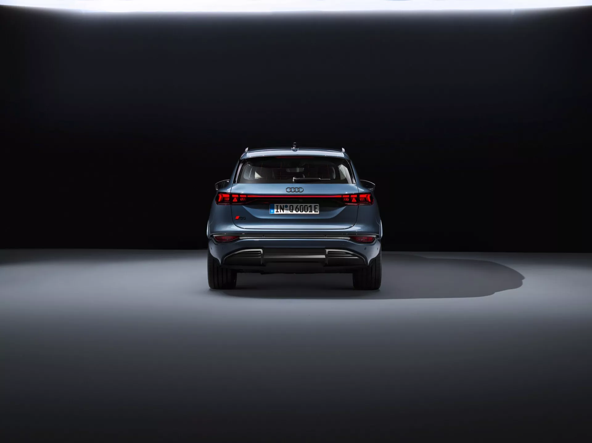 Bakre vy av Audi Q6 e-tron 2024 mot en mörk bakgrund som visar dess eleganta design, centrala emblem, modellnamn, horisontell ljuslinje, bakre spoiler och diffuser-liknande stötfångare med reflekterande element.