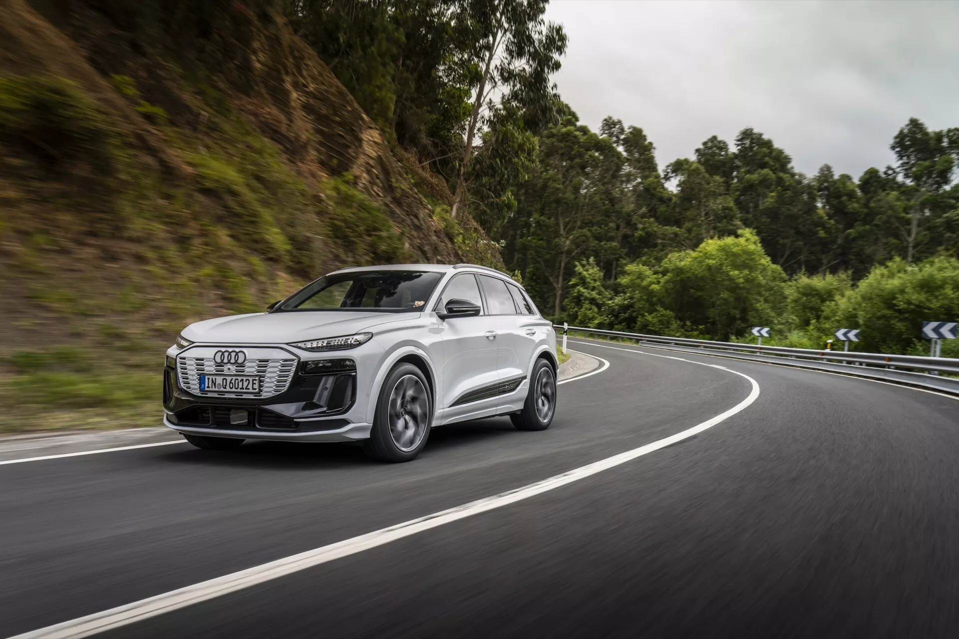 Bilden fångar en dynamisk sidovy på en modern vit SUV, sannolikt en Audi Q6 e-tron 2024-modell, som färdas på en väg omgiven av ett skogsområde. Fokus ligger på fordonets frongrill, strålkastare, sidoprofil, hjul och sidobackspeglarnas aerodynamik. Strålkastarna är eleganta med LED-teknik, vilket antyder avancerad belysning. Fälgarna i lättmetall med ett flerspokesdesign antyder högprestandaegenskaper. Bilens rörelse på en kurvig väg framhäver dess hanteringsförmåga och eventuellt avancerade förarstödsystem. Den suddiga bakgrunden betonar hastighet och effektivitet vid navigering genom utmanande scenarier.