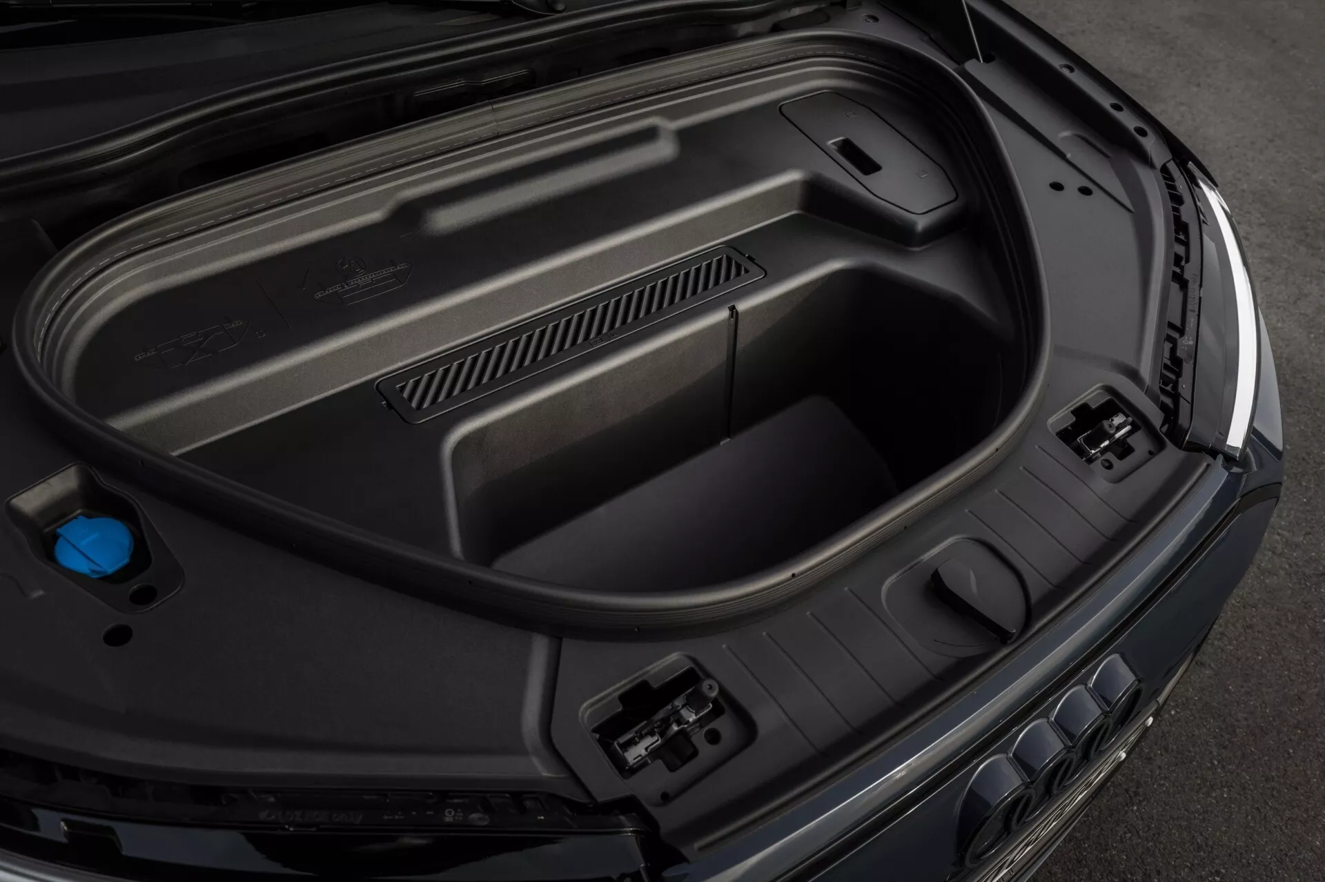 Närbild från hög vinkel av Audi Q6-e-Tron 2024s bakre bagageutrymme, som visar interiöregenskaper inklusive formgjutna plastpaneler för hållbarhet, strömtillbehörsuttag på höger sida, inbäddat förvaringsutrymme i botten och en blå kork på vänster sida, vilket indikerar avancerad bilteknologi och en elegant, premium finish.