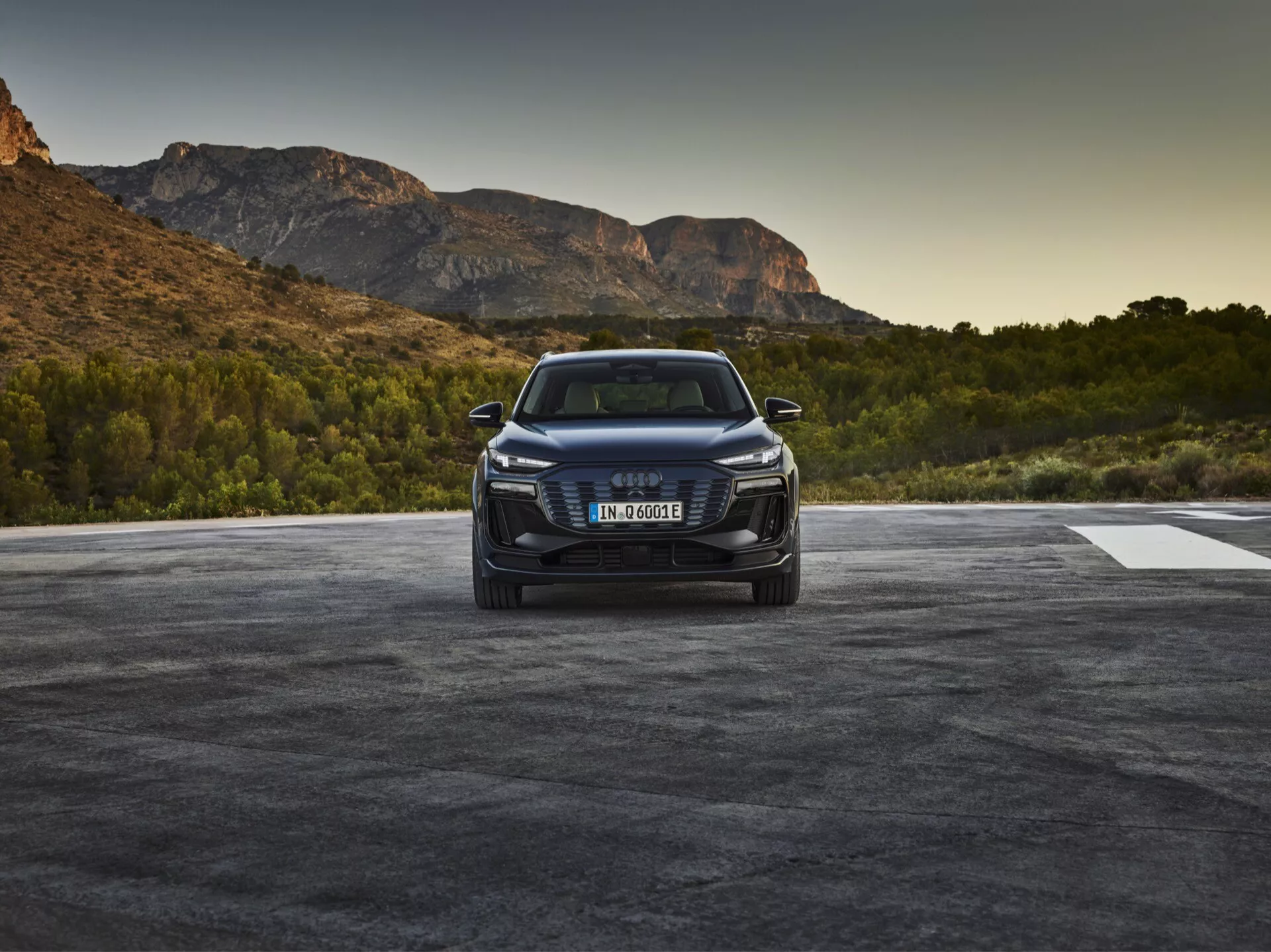 Bild av en Audi Q6 e-tron 2024 fotograferad från en trekvarts framvinkel, som visar fordonets främre grill med varumärkets emblem, skarpa LED- eller laserstrålkastare och detaljerad främre stötfångare med luftintag och möjligen sensorer eller kameror som indikerar förarassistansfunktioner. Motorhuven har konturlinjer och den eleganta sidoprofilen tyder på aerodynamik, med en högre SUV-ställning. Bilen är placerad i en utomhusmiljö med naturligt landskap som antyder soluppgång eller solnedgång.