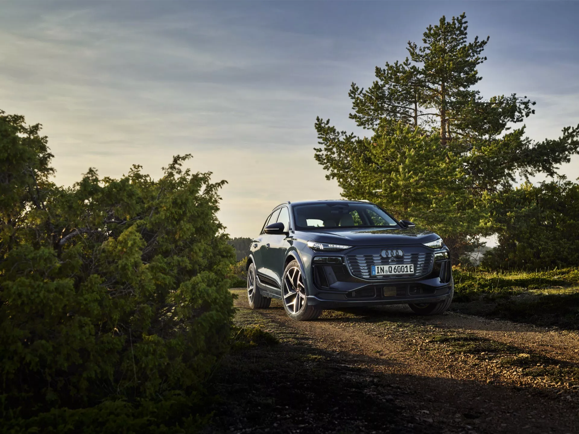Trekvarters framåtvänd vy av Audi Q6 e-tron 2024 parkerad utomhus på en grusstig mitt i naturen, fotograferad i varm, gyllene morgon- eller eftermiddagsljus. Den upphöjda vinkeln visar den djärva frontgrillen med ett centrerat emblem, eleganta strålkastare som antyder avancerad LED- eller Matrix LED-teknologi och utmärkande sportiga lättmetallfälgar. Fotot inkluderar naturliga element i bakgrunden, vilket indikerar bilens mångsidiga kapacitet både i stadsmiljö och vid lättare terrängaktiviteter.