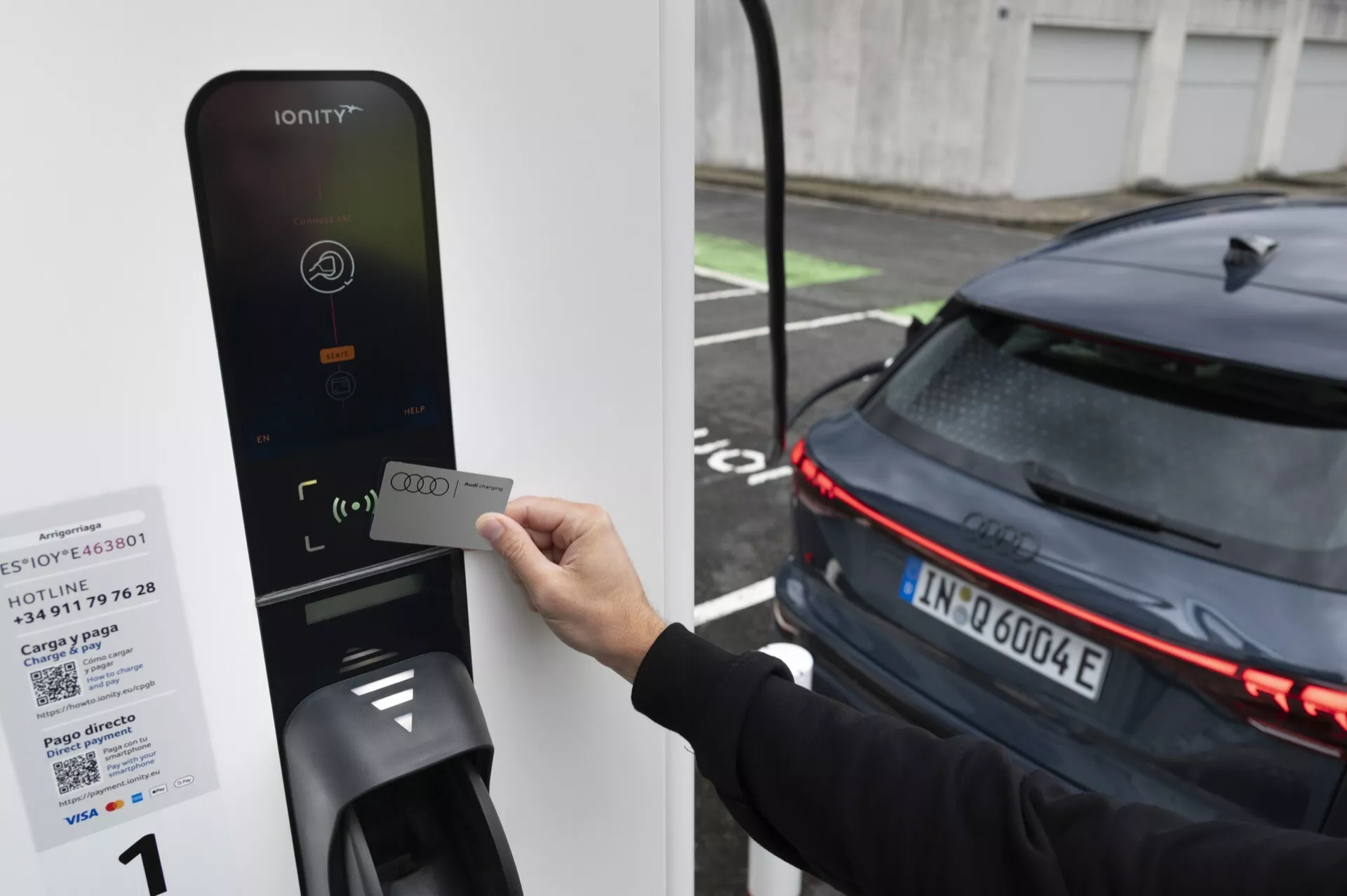 Bild av en person som håller ett RFID-kort nära en IONITY-laddstation för elfordon, riktat mot bakre delen av en modern, mörk bil med horisontella LED-bakljus, vilket indikerar att den är elektrisk. Bilen, förmodligen en EV, är parkerad bredvid laddaren, vilket antyder att den laddas eller förbereds för laddning.