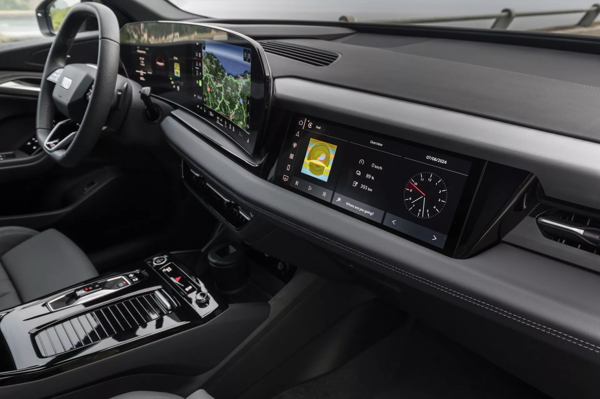 Interiörvy av 2024 Audi Q6 e-tron med fokus på instrumentbrädan och mittkonsolen från en något förhöjd vinkel. På vänster sida syns ratten, vilket indikerar vänsterstyrd konfiguration, medan mittkonsolen innehåller en växelspak, olika kontroller och en stor pekskärm. Den digitala instrumentpanelen visar kördata. Den eleganta instrumentbrädan sträcker sig horisontellt med ventilationsöppningar och ett annat designelement på höger sida. Avancerad teknik framgår med den centrala pekskärmen som visar navigations- och multimedia information, en heads-up display synlig ovanför ratten och touch-känsliga knappar för klimatkontroll. Designen betonar minimalism och premium material, med lädersömmar och metall accenter som förstärker den lyxiga känslan.