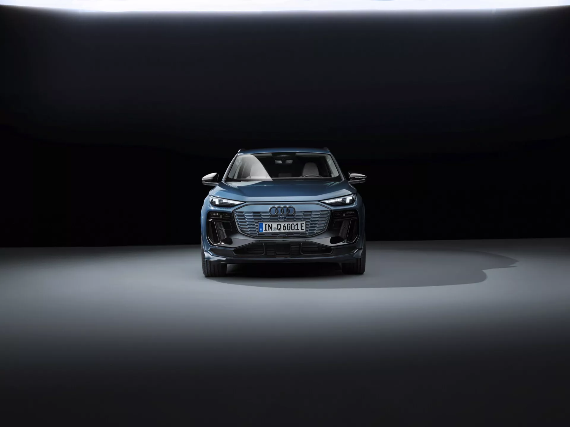 Främre vinkelvy av 2024 Audi Q6 e-tron som visar huven, grillen, delar av taket och frontdesignen. De eleganta strålkastarna har sannolikt LED-teknik. Den tyska registreringsskylten antyder att det är en elektrisk eller hybridmodell. Karakteristisk Audi Singleframe-grill, ytterligare intag och potentiella säkerhetssensorer, som alla betonar dess avancerade förarassistanssystem. Det dramatiska ljuset med mörk bakgrund och belysning ovanifrån framhäver bilens lyxiga och sofistikerade design.