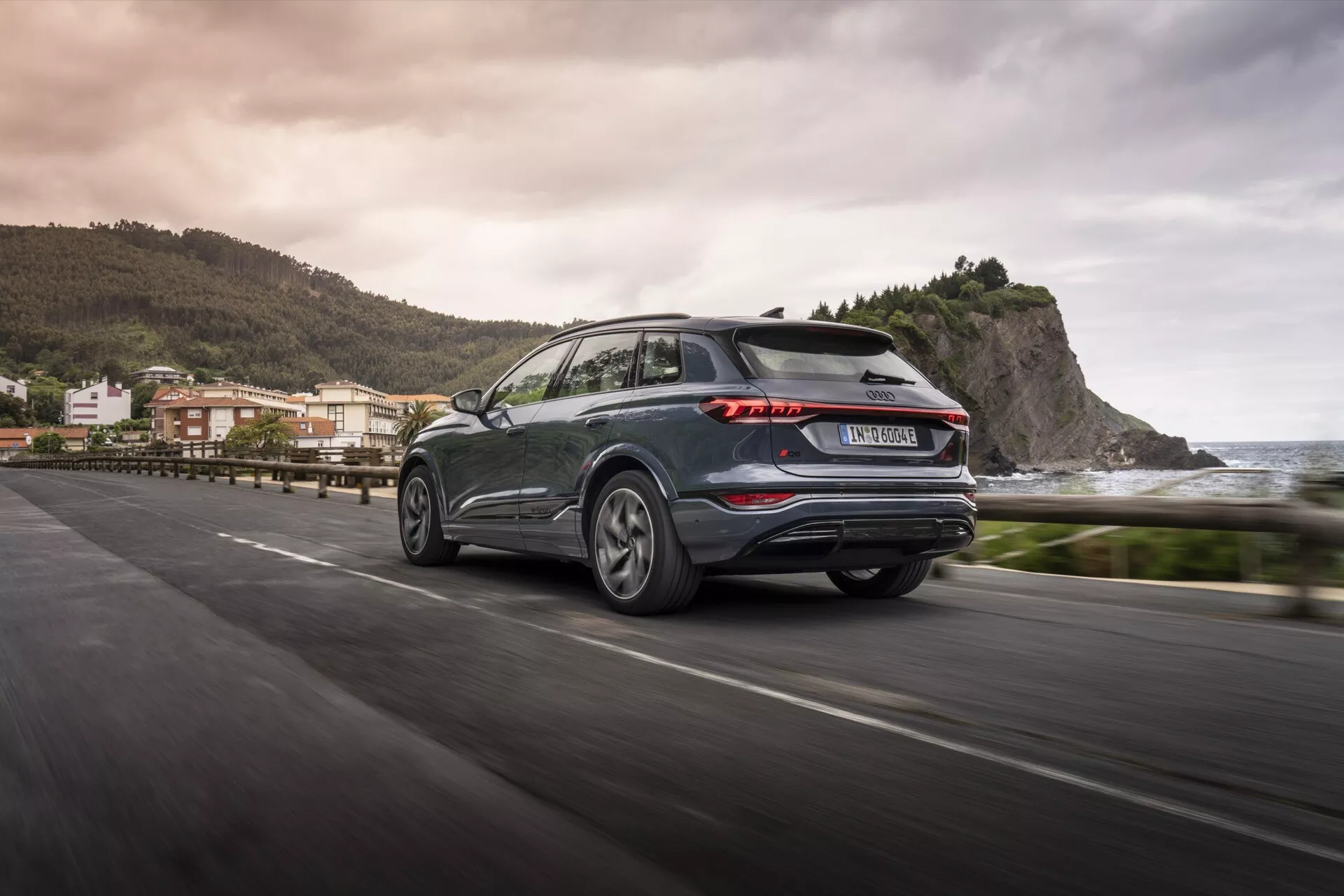 Audi Q6 e-tron 2024 bakre trekvarts vy i rörelse på en kustväg, som visar karakteristiska LED-bakljus, mörk metallic färg, lättmetallfälgar och praktisk bakrutetorkare. Den suddiga bakgrunden antyder en kustmiljö, som reflekterar en modern SUV:s estetik och teknologiska framsteg.