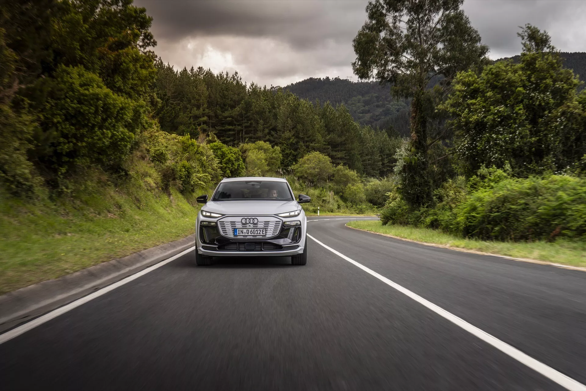 Audi Q6-e-tron 2024 fångad i rörelse från en frontvy på en tvåfilig väg omgiven av grönska. Bilden visar fordonets grill, strålkastare, motorhuv och främre stötfångare, och framhäver dess dynamiska prestanda och design. Bilen har distinkta LED-strålkastare, ett framträdande lyxemblem på grillen och aerodynamiska linjer, allt under molnigt väder med torra vägförhållanden.