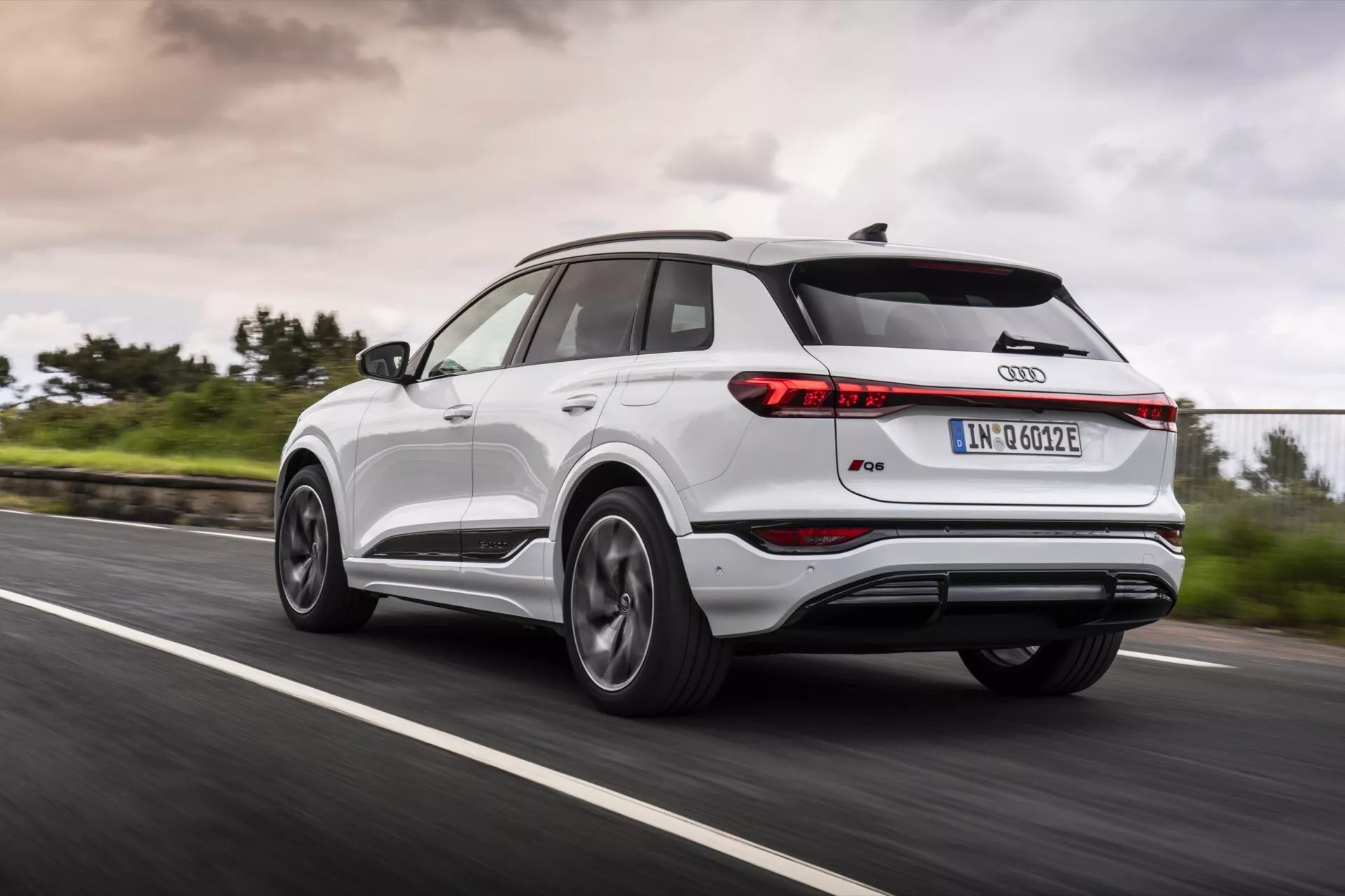 Audi Q4 e-tron 2024 som kör på en asfalterad väg, visad i trekvartsvy bakifrån och fångar vänster bakre sida. Bilden visar smala bakljus, takspoiler, panoramasoltak, multiekrade aluminiumfälgar, tvåfärgad kaross med metalltrim och den ikoniska Audi-ringsymbolen. Rörelseoskärpa på hjulen och grönska vid vägkanten med molnig himmel antyder dynamiska körförhållanden för alla väderförhållanden.