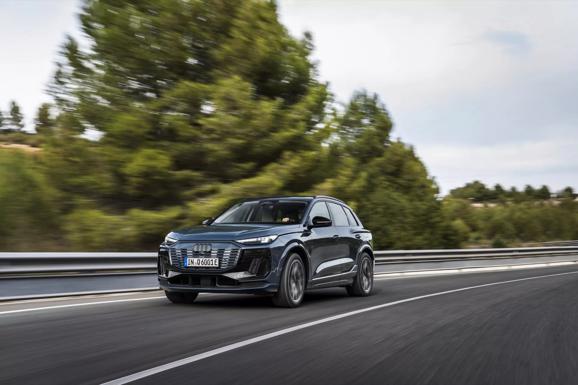 Audi Q6 e-tron 2024 SUV i rörelse på en motorväg, fångad från en trekvarts frontvy. Bilden visar framgrill, LED-strålkastare, huven, vindrutan och en del av sidoprofilen, inklusive dörrar och sidospeglar. Fordonet kör på höger sida av körfältet, och rör sig från vänster till höger med en suddig bakgrund som indikerar hastighet. Träd och ett räcke är svagt synliga. Den tydliga designen av framstrålkastarna antyder avancerade belysningssystem, och den stora grillen antyder kylteknik eller sensorer för ADAS. Sidospeglar kan innehålla blinkers eller döda vinkeln-övervakningssystem. Hjul ger insikt i prestanda eller lyxkategorier.