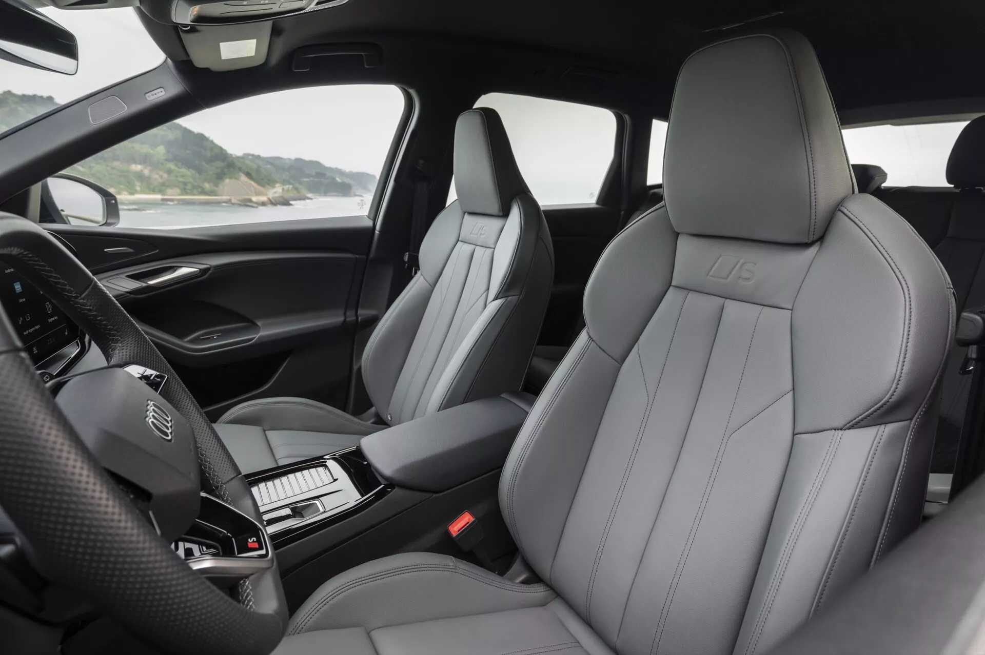 Interiörvy av Audi Q6 e-Tron 2024 som visar lyxiga lädersäten fram med framträdande sömnadsmönster, präglad logotyp och modern design. Fotografin framhäver instrumentpanelen med en ratt med monterade kontroller och ett oupplyst digitalt instrumentkluster. Mittkonsolen som hyser växelspaken och prydligt arrangerade knappar är delvis synlig. Bilden fångar de vadderade dörrarmstöden med kontroller för speglar, fönster och möjligen justeringar av säten. Det naturliga ljuset framhäver kupéns komfort och högkvalitativa material.