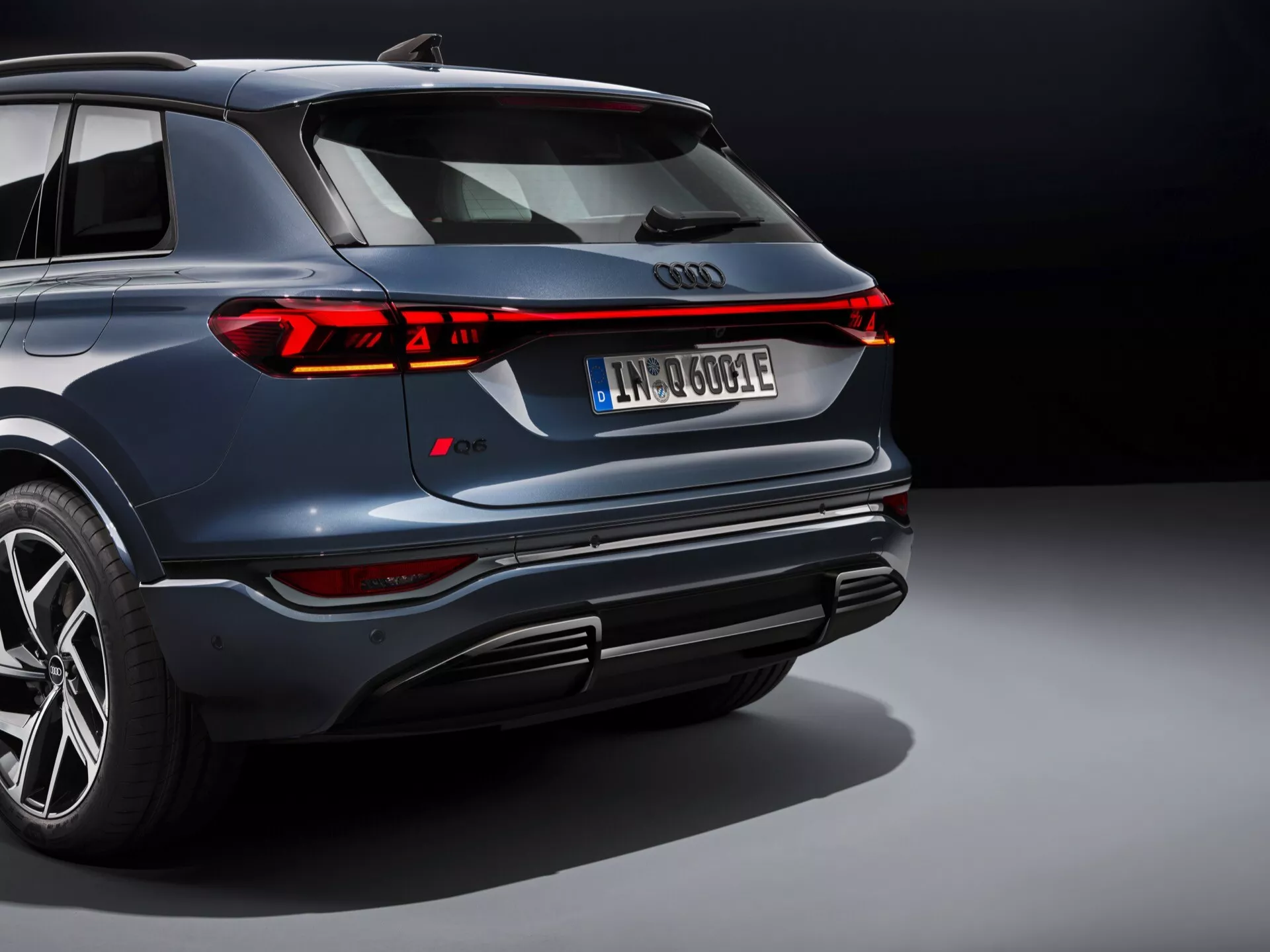 Den här bilden visar en modern SUV, sannolikt Audi Q6 e-tron 2024, sett från en upphöjd bakvinkel. Den ikoniska fyraringade Audi-emblemet syns tydligt bakpå, omgivet av en elegant kontinuerlig LED-bakljusremsa som sträcker sig över fordonets hela bredd, med segmenterade bromsljus och blinkers. Den mörkt marinblå bilen har en polerad metallisk finish, en djärv stötfångardesign med integrerade avgaser och diffuser, flerfälgade lättmetallfälgar med lågprofildäck och en registreringsskylt som antyder en elektrisk eller hybrid drivlina. Det professionella ljuset och den neutrala bakgrunden framhäver fordonets lyxiga och avancerade design och betonar dess sportiga och högklassiga attribut.