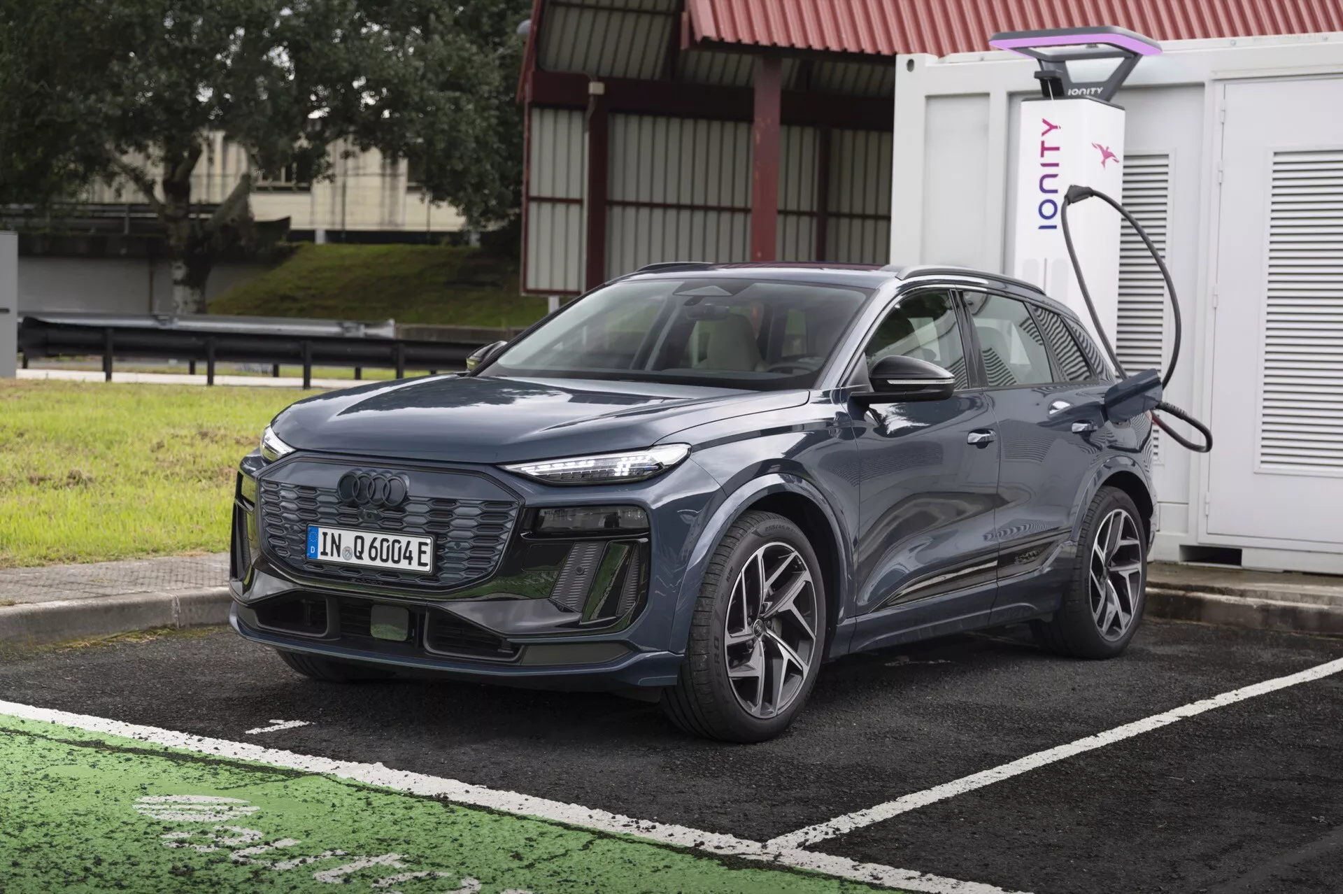 Bilden visar en modern Audi Q6 e-Tron 2024 SUV parkerad vid en laddningsstation för elfordon. Bilen visas från en något vinklad framvy, vilket framhäver dess grill, strålkastare och en del av huven och vindrutan. Den är placerad diagonalt med fronten något vänd till vänster. Den vänstra sidan av fordonet, inklusive framhjulet på förarsidan, dörren och sidospegeln, är synlig. SUV:en är ansluten till en laddningsstation på den vänstra sidan framtill ovanför hjulet. Bilen har eleganta linjer, en stängd grill som är vanlig för elbilar och avancerade LED-strålkastare. Detaljerna antyder möjliga avancerade förarassistanssystem som adaptiv farthållare, filhållningsassistent och kollisionsundvikande system. Bilden är ljus och klar, tagen under dagen utan rörelseoskärpa, vilket gör alla design- och teknikelement tydligt synliga.