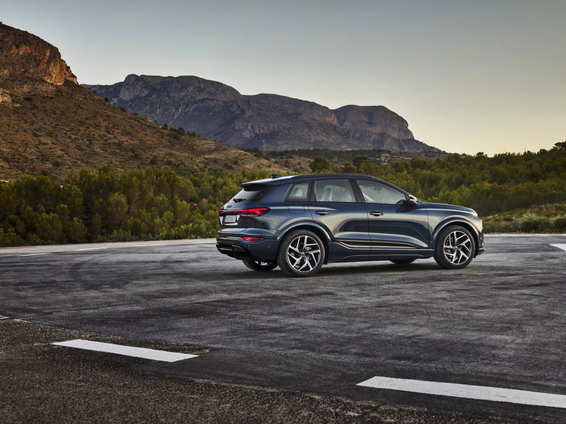 Bild av en 2024 Audi Q6-e-tron SUV parkerad på en utomhusparkering med ett naturligt, möjligtvis bergigt landskap i bakgrunden. Fotot fångar en trekvarts bakvy, som framhäver både sidans och bakdelens designelement. Förarsidan är tydligt synlig och visar designlinjer, bakrutor och hjuldesign. Den sluttande taklinjen antyder en sportig, coupéliknande SUV-design. De moderna LED-bakljusen, synliga sensorer eller reflektorer på bakre stötfångaren och ett tvåtonssystem framhäver höjden och hållningen. Stora aluminiumfälgar och rejäla däck indikerar en högre utrustningsnivå med sportig estetik. Fordonet verkar vara nära kameran, vilket ger en detaljerad vy. Anmärkningsvärda funktioner inkluderar LED-belysning, parkeringssensorer, högpresterande hjul och däck, aerodynamisk styling och troligen avancerade förarassistanssystem och fyrhjulsdrift.