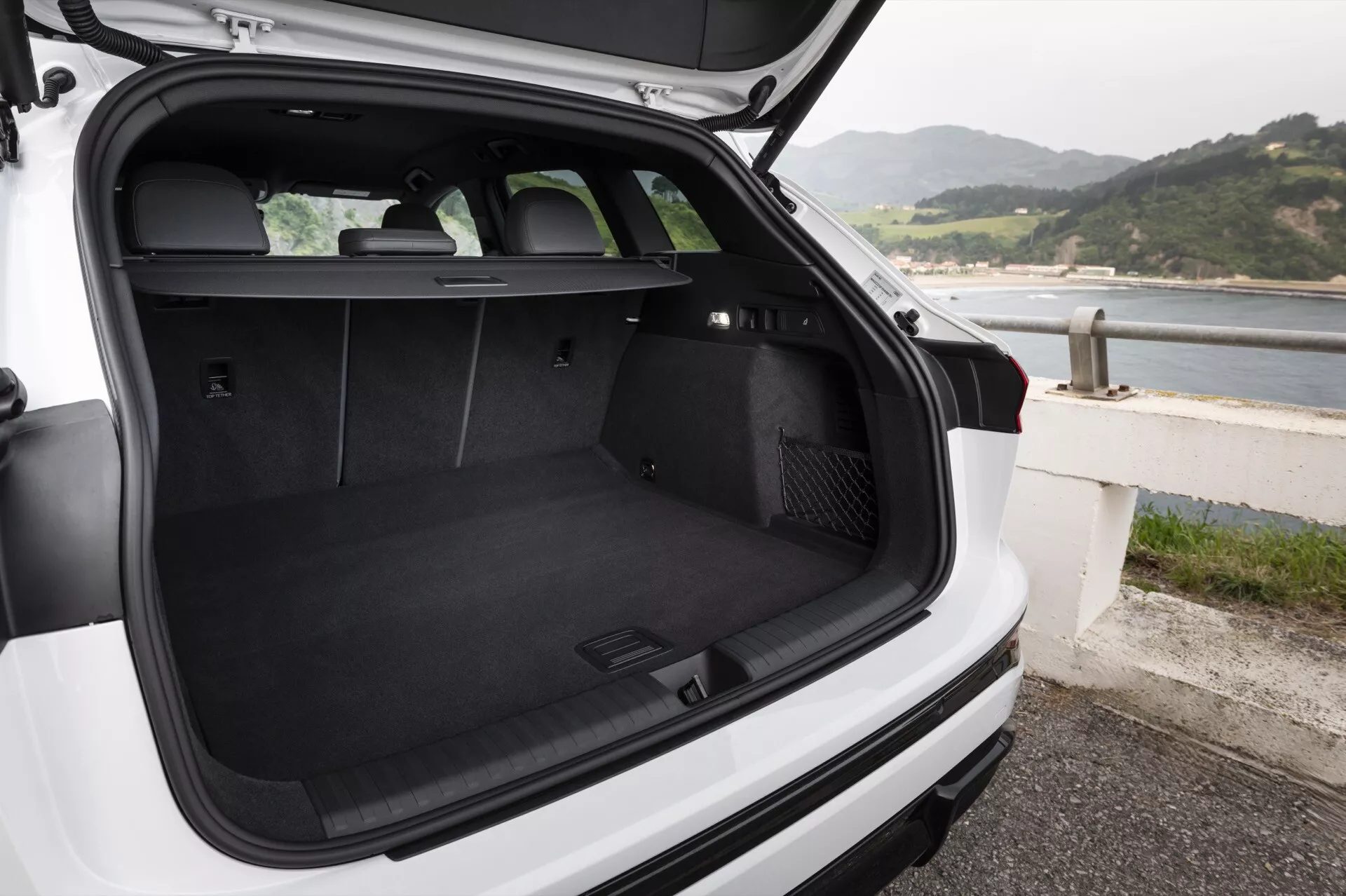 Bilden visar lastutrymmet på en vit Audi Q6 e-Tron 2024 hatchback eller SUV, sett bakifrån med öppen baklucka och en något neråtriktad kameravinkel. Den svart-mattade bagageutrymmet är tomt och visar dess rymlighet och rena skick. Baksätena är uppfällda, vilket indikerar möjlighet att transportera passagerare eller fälla ner sätena för mer lastutrymme. Ett infällbart lastskydd är placerat strax bakom baksätena men är inte utfällt, vilket ger en fullständig vy över bagageutrymmet. På vänster sida av lastutrymmet finns ett 12-voltsuttag och på höger sida verkar det finnas ett fack, troligtvis för förvaring av små föremål. Den bakre vindrutetorkaren är synlig. Fordonet är parkerat nära en kustlinje, vilket antyder en havsnära plats. Det finns inga föremål eller personer i bagageutrymmet.