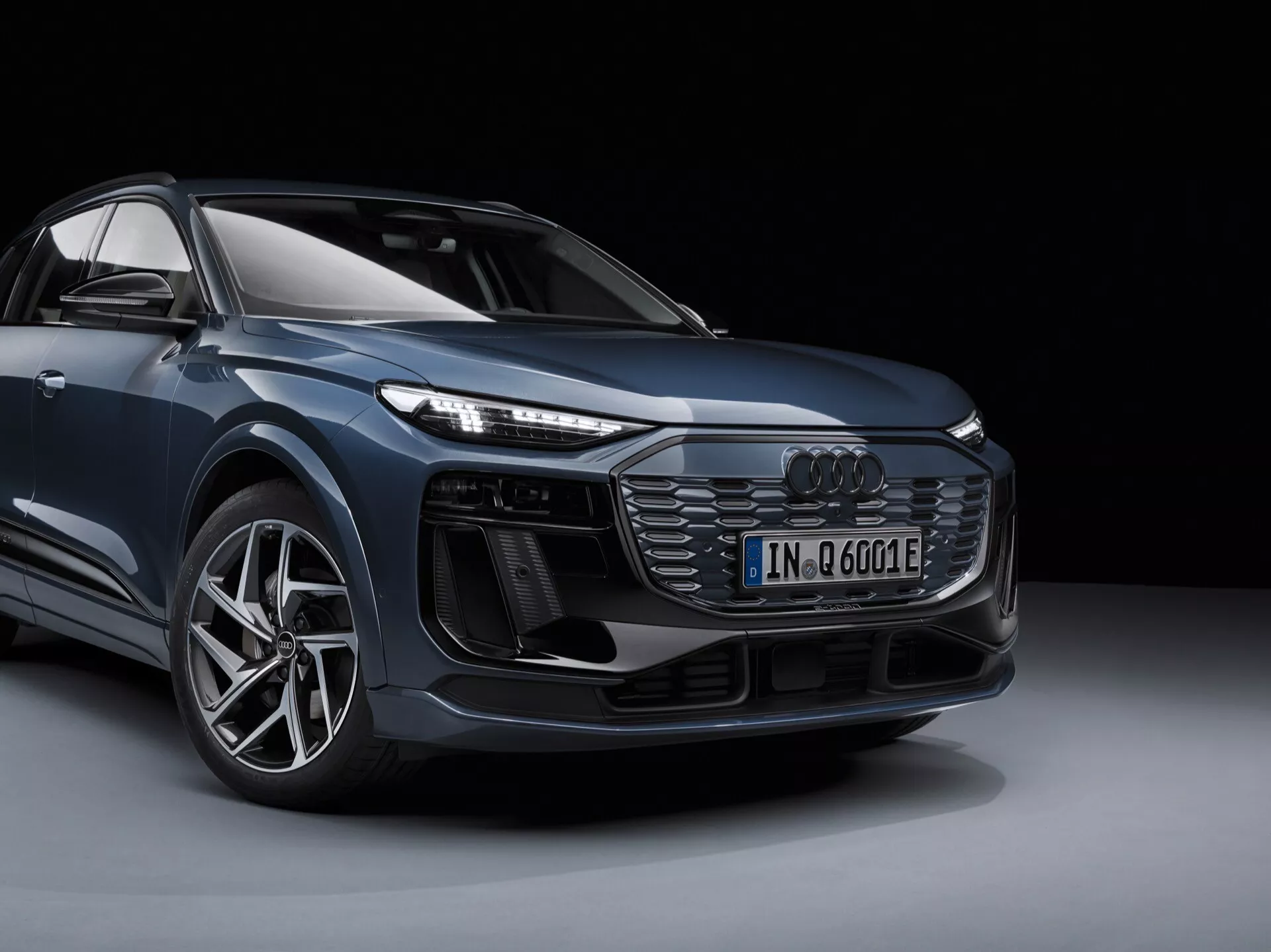 Bilden visar en modern Audi Q6 e-tron 2024, fångad från låg framvinkel i trekvarts vy. Synliga är den ikoniska frontgrillen med horisontella slats, intrikata LED-strålkastare, aggressiv främre stötfångare med stora luftintag, mångekrade aluminiumhjul och emblem för den elektrifierade modellen. Den eleganta och sofistikerade SUV:en visar upp aerodynamiska linjer och avancerad design i en professionell studiosession mot svart bakgrund.