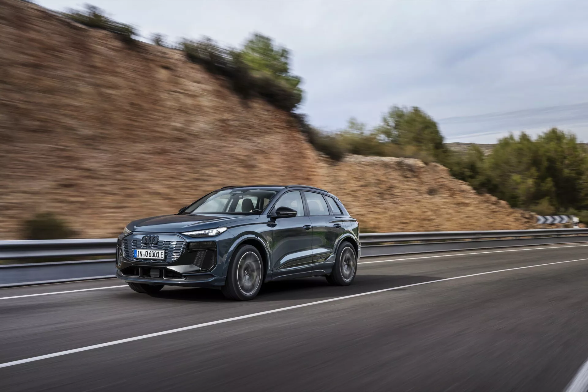 Denna bild visar en elegant och sofistikerad Audi Q6 e-tron 2024 SUV, fångad från en sidovinkel medan den är i rörelse. Fordonets moderna design framhävs av dess anmärkningsvärda LED-strålkastare och aerodynamiska egenskaper, inklusive en sluttande taklinje och strömlinjeformade sidospeglar. Bilen, i en mörk färg, troligtvis grå eller svart, kör på en högfartsväg, vilket framgår av den suddiga bakgrunden som skapats genom en panoramafotograferingsteknik. Framhjulet är lätt vridet mot kameran och visar designen av lättmetallfälgen. Ytterligare synliga element inkluderar möjlig integrerad teknologi i strålkastarna och sidospeglarna samt potentiella takrelingar med fokus på funktionalitet.