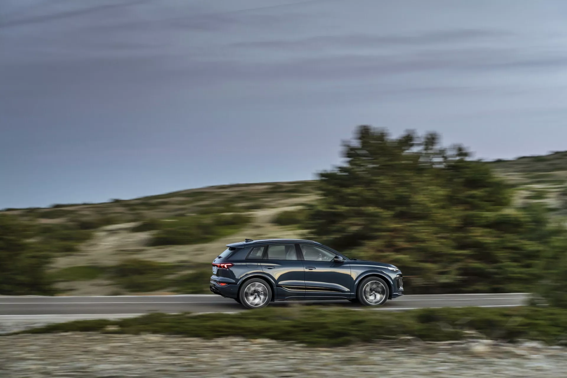 Dynamisk sidovy av Audi Q6 e-tron 2024 i rörelse med oskarp bakgrund, medelavstånd parallell vinkel som visar profil, designdetaljer, fönster, fem dörrar, sidospeglar, fälgar i legering, dagsljusinställning, lantlig bakgrund.