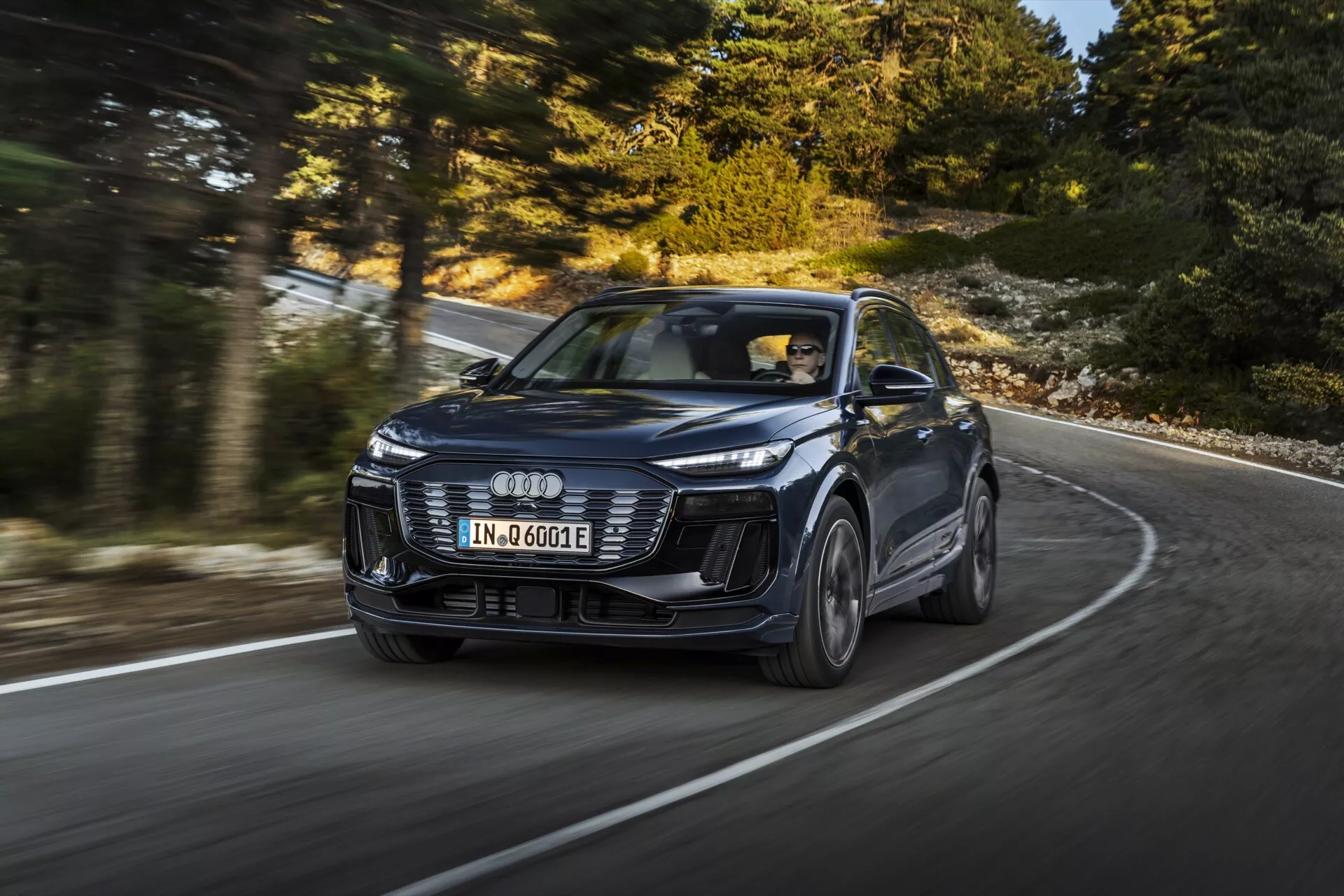 Bild av en modern SUV Audi Q6 e-tron 2024 i rörelse, fångad från en dynamisk trekvartsvinkel fram som framhäver dess design och prestanda. Fordonet, identifierat av den distinkta fyraringade logotypen, har emblematiska LED-strålkastare, en aggressiv främre stötfångare och skulpterade motorhuvslinjer. Bilden, tagen från en lägre vinkel, framhäver bilens närvaro på en svängd väg, med lätt suddade hjul som indikerar rörelse. Den nya modellen föreslår avancerade teknologier som adaptiv farthållare och elektrisk drivning, markerad med 'e-tron'-emblemet. Professionell fotosession som visar lyx, design och teknologi i vänsterstyrd konfiguration.