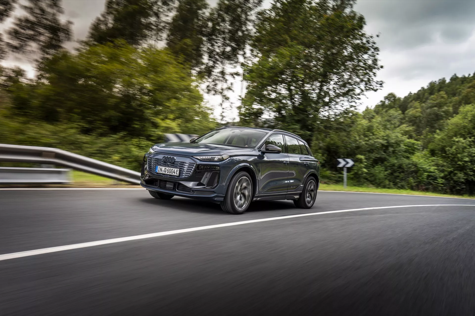 Bilden visar en modern Audi Q6 e-tron 2024 SUV i rörelse, fångad från en sida-fram-vinkel, med fokus på dess vänstra sida. Den dynamiska bilden framhäver den framträdande vänstra strålkastaren med distinkta LED-dagljus, ett stort samtida galler, eleganta aerodynamiska sidobackspeglar med integrerade blinkers och stora legeringsfälgar med lågprofildäck. Bilen har ett panoramasoltak och tonade rutor, med en reflekterande metallisk lack. Den låga bildvinkeln och den lätta suddigheten i bakgrunden betonar bilens hastighet och rörelse på en kurvig tvåfilig väg.