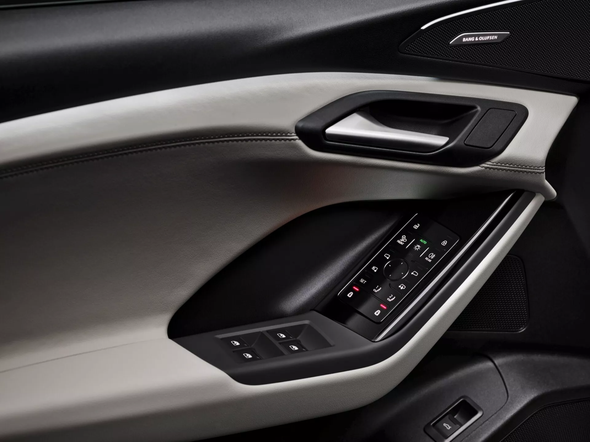 Närbild av dörrpanelen på insidan av Audi Q6 e-tron 2024, som visar designfunktioner och detaljer. Dörrpanelen, fotograferad från en diagonal vinkel, avslöjar olika material och texturer, inklusive en mjuk övre sektion med synliga sömmar, en hård trim som rymmer ett ergonomiskt dörrhandtag och en vadderad armstöd med bakgrundsbelyst kontrollpanel för fönster och justering av sidobackspeglar. Under armstödet finns ett märkesljudsystem som antyder ett premiumljudsystem. Färgskalan involverar en tvåtonad design som förbättrar estetiska och funktionella kvaliteter.