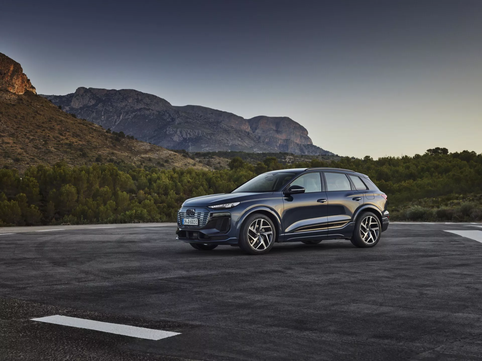 Tre fjärdedels frontvy av en blå Audi Q6 e-tron SUV 2024 parkerad utomhus, med framträdande grill, stiliga strålkastare, skulpterad huva, stora lättmetallfälgar, sidospeglar och dörrar på förarsidan mot en naturskön bergsutsikt i tidigt morgon- eller sent eftermiddagsljus.