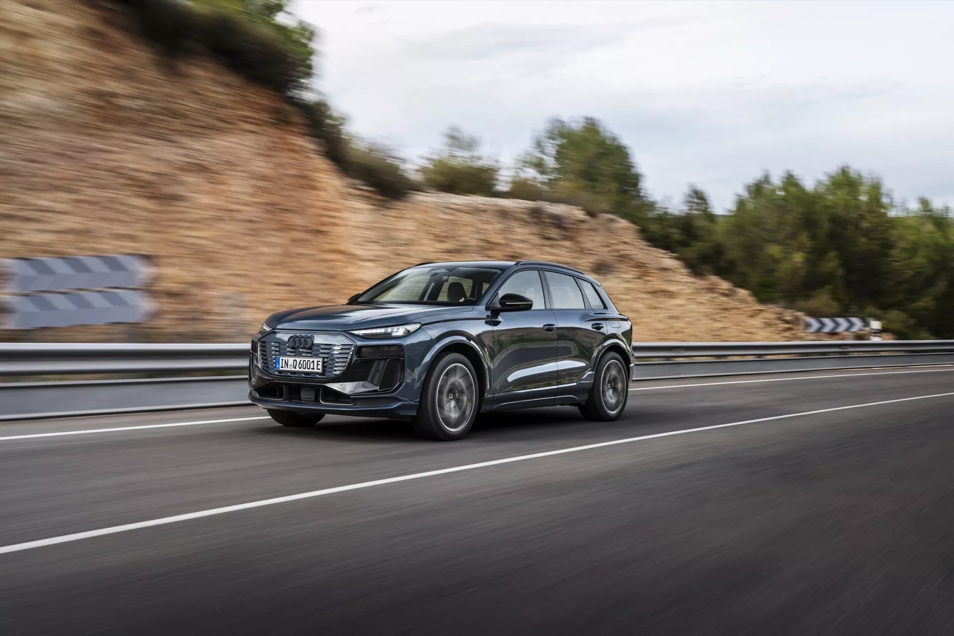 Bild av 2024 års Audi Q6 e-tron i rörelse, fångad med hjälp av en panoreringsmetod för att framhäva SUV:ns eleganta profil och moderna design. Bilen, inramad från sidan, susar fram på en flervägs motorväg med suddiga hjul och bakgrund, vilket understryker dess dynamiska kapacitet. Det mörka metalliska fordonet har en stor främre grill, distinkta LED-strålkastare, sportiga aluminiumhjul och reflekterande ytor. Den molniga himlen och den blöta vägen skapar en dramatisk scen, som framhäver bilens lyx- och prestandafunktioner.