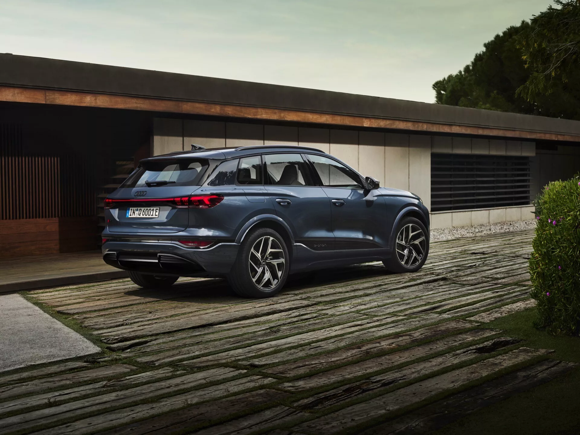 Bild av 2024 Audi Q6-e-Tron parkerad på ett trädäck, som visar en trekvarts bakvy. Den lyxiga SUVen har en synlig sidoprofil och bakdel med moderna horisontella LED-bakljus, en robust baklucka, en framträdande bakre stötfångare med möjliga parkeringssensorer, och en diffusor under med dubbla avgasrör. Sidoprofilen framhäver stora, flerspakiga lättmetallfälgar, distinkta karaktärslinjer, takräcken och något tonade bakrutor. Teknologiska tillägg som sidospeglar med integrerade blinkers och infällda dörrhandtag antyder ett avancerat, högkvalitativt fordon i en sofistikerad, minimalistisk miljö.