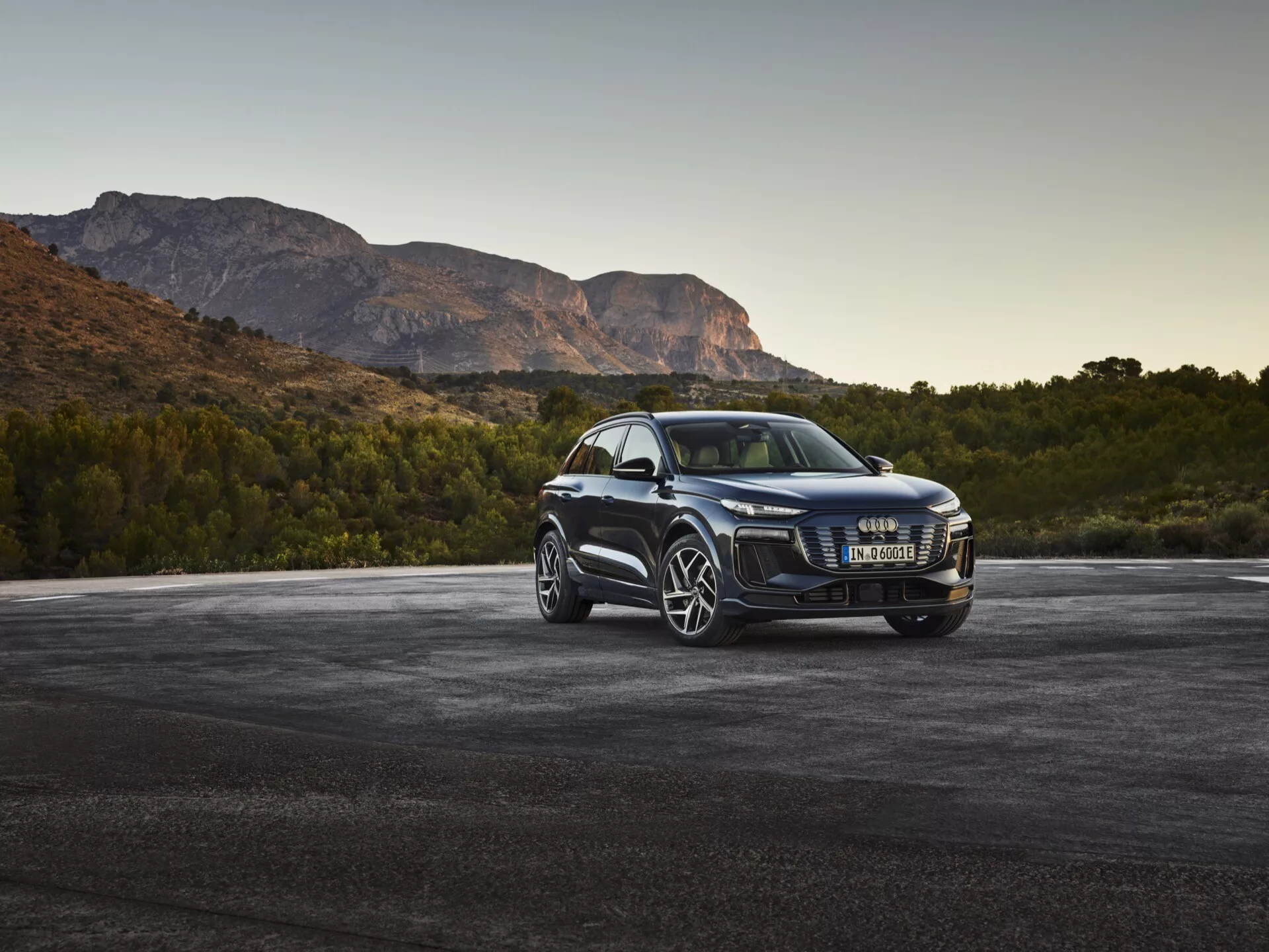 2024 Audi Q6 e-tron lyx-SUV i trekvart vinkling framifrån som visar kromförsedd grill, moderna LED-strålkastare, stora flerspakiga fälgar, sidospeglar med integrerade blinkers, tonade rutor och glansig lack, parkerad på asfalt med berg i bakgrunden vid gryning eller skymning.