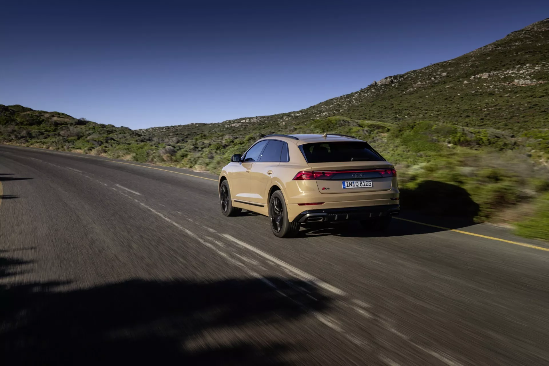 Audi Q8 2023 (4M-Facelift) i rörelse, fångad från en bakre trekvartsutsikt som visar höger sida och bakdel på en delad motorväg med naturligt kulligt bakgrund under blå himmel, med en kupéliknande taklinje, elegant design, förlängda bakljus, ljusbeige lack och dubbla avgassystem.