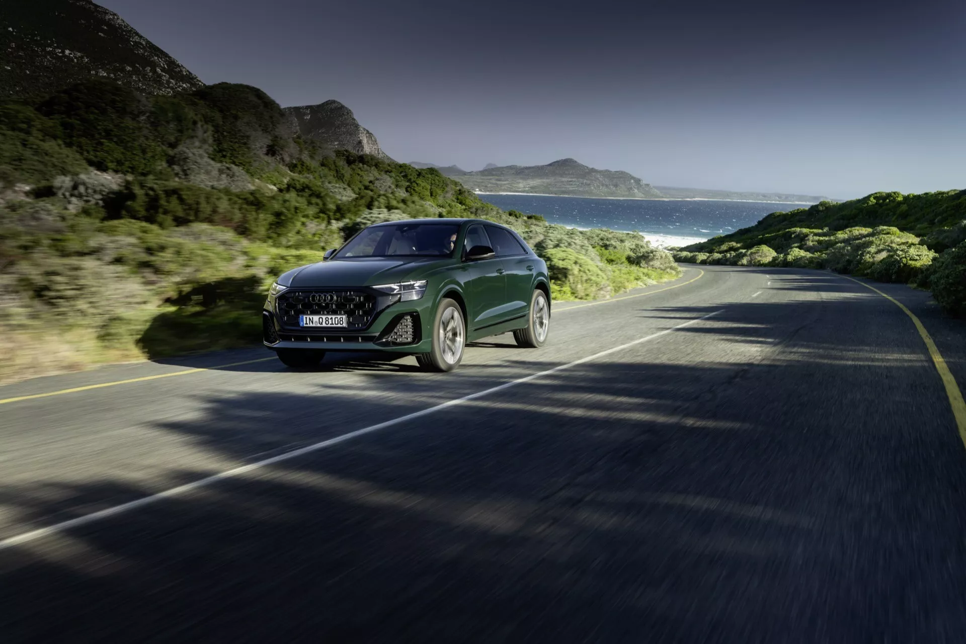 Audi Q8 2023 (4M-Facelift) kör på en kustväg, med en front-sidovy som visar dess stora grill, LED-strålkastare, slanka motorhuv och del av sidopanelen, inneslutande teknik och lyx i rörelse mot en bakgrund av klar himmel, berg och ocean.
