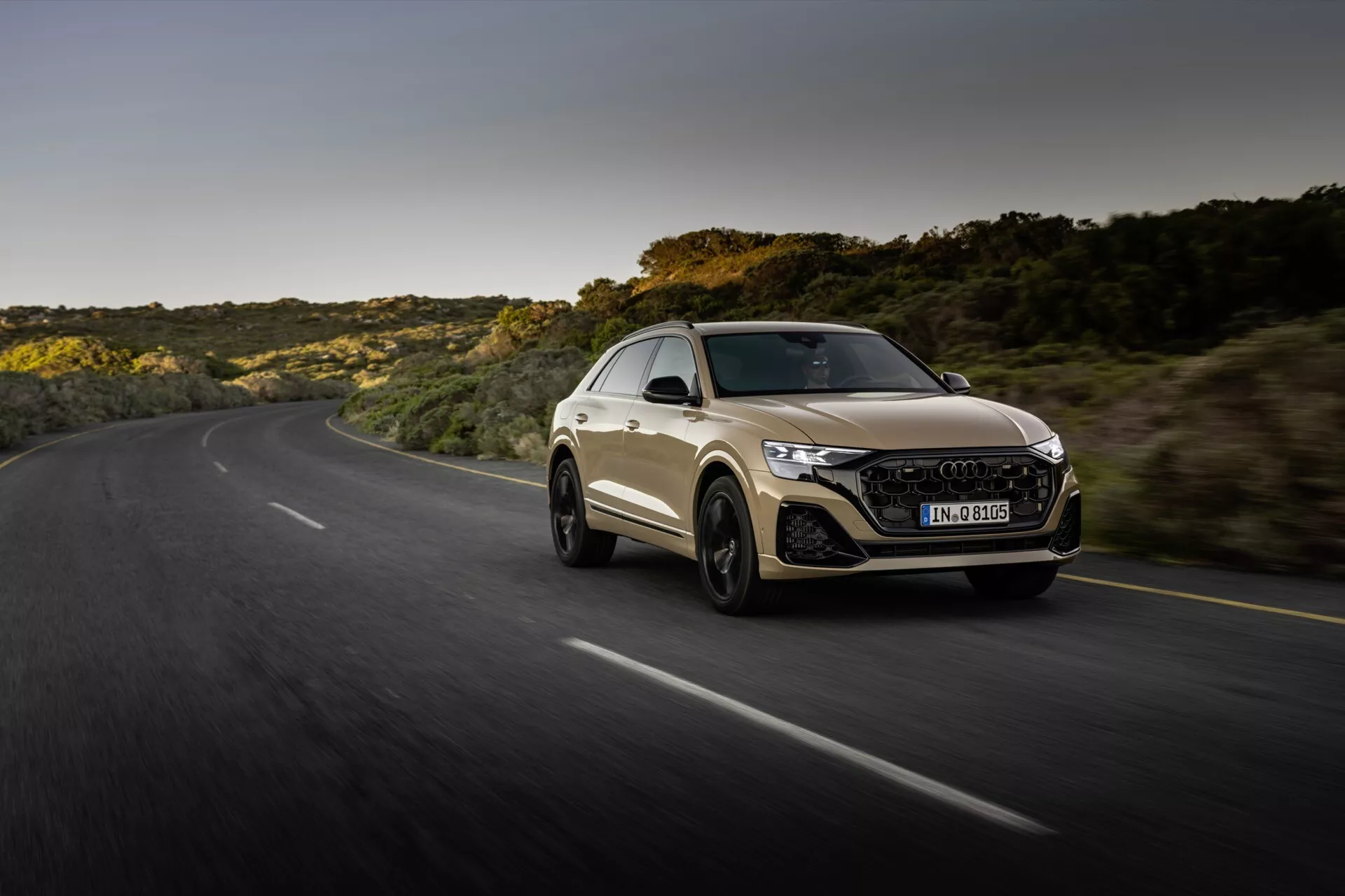 Audi Q8 2023 (4M-Facelift) i en dynamisk körning på en slingrande väg under gyllene timmen, visar bilens sportiga coupédesign, aggressiv front med LED-strålkastare och hexagonal grill, och aerodynamisk silhuett mot en naturlig landskapsbakgrund.