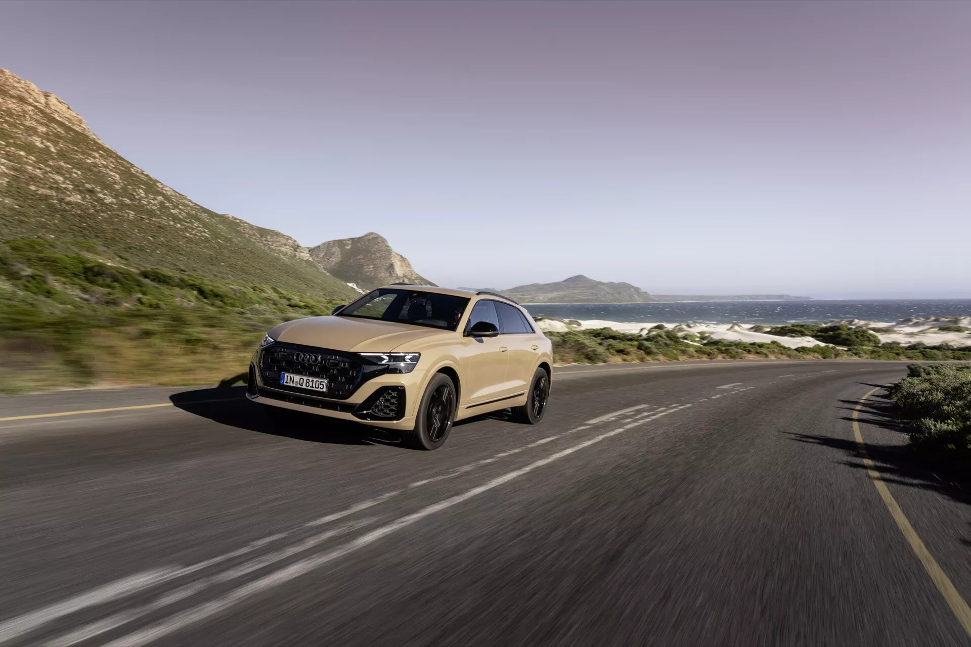 Trekvarts framifrån vy av Audi Q8 2023 (4M-Facelift) på en kustväg, som visar LED-strålkastare, aerodynamisk design och rörelseoskärpa som indikerar rörelse