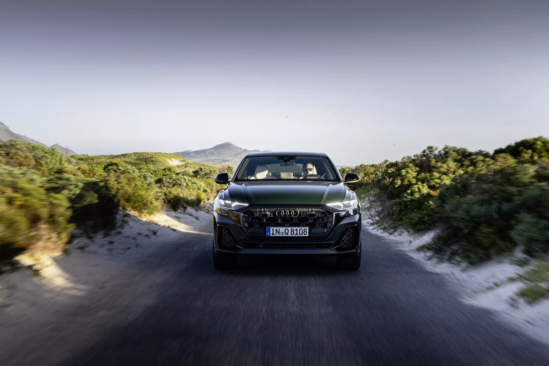 Audi Q8 4M-Facelift 2023 i rörelse, frontvy som visar LED-strålkastare, avancerad grilldesign och aerodynamiska funktioner mot en dynamisk väg- och bergsbakgrund