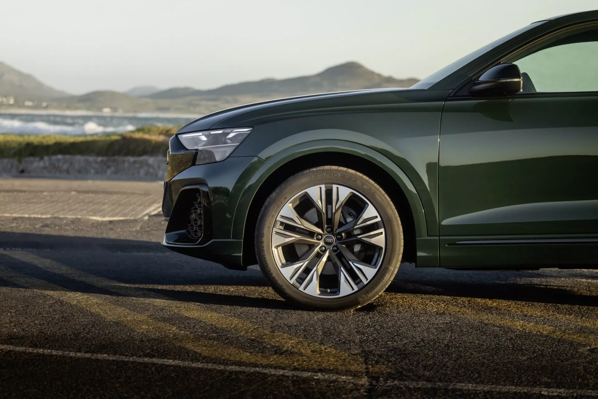 Närbild av 2023 Audi Q8s (4M-Facelift) sportiga främre vänstra kvartal, inklusive det polerade, flerspakiga legeringshjulet, delvis frontstötfångare med stort luftintag, kant av modern LED-strålkastare och glänsande djupgrön metallisk lack på karossen, mot en utomhusbakgrund.
