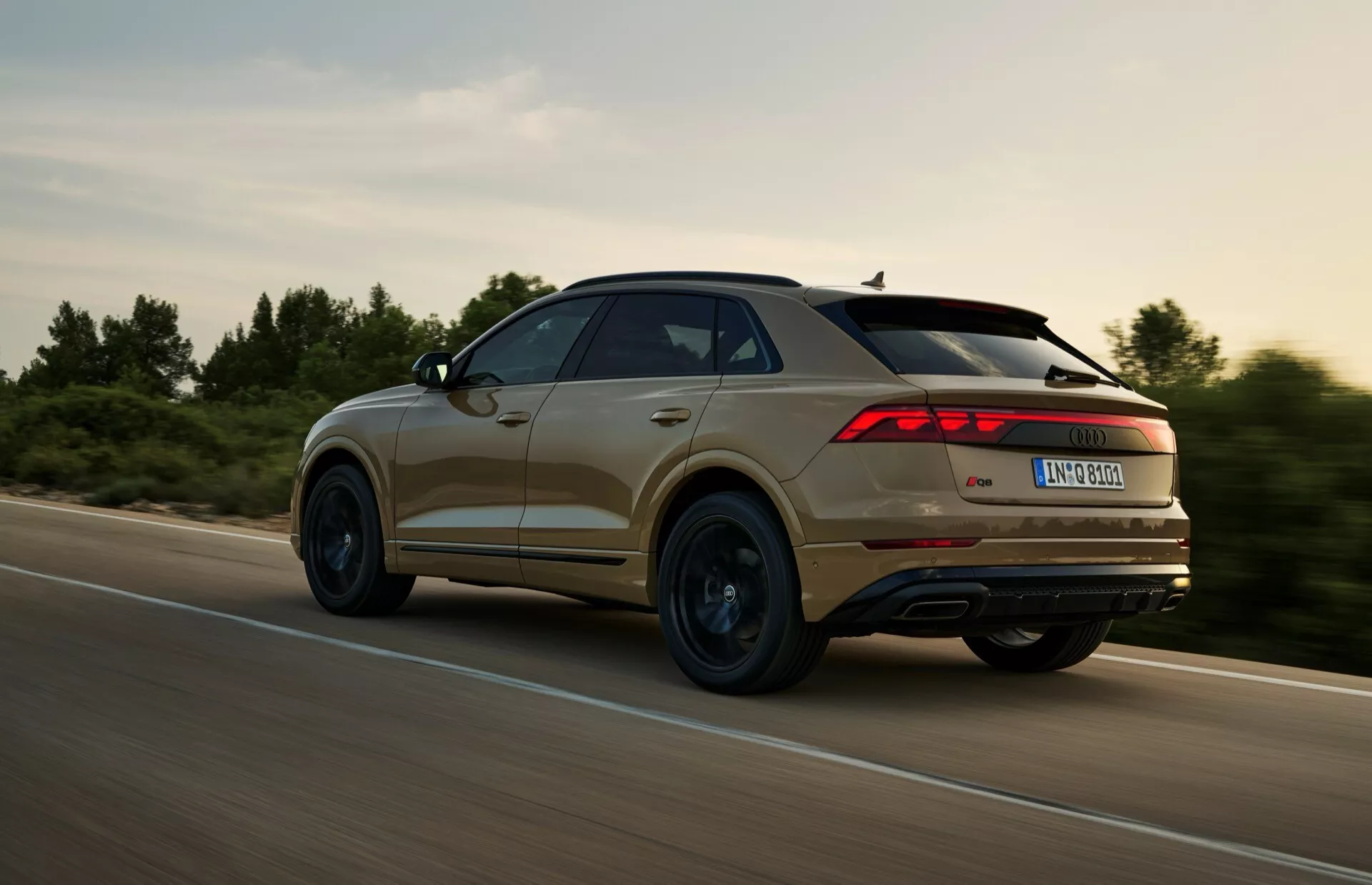 Bakre tre fjärdedelsvy av en SUV Audi Q8 (4M-Facelift) 2023 i rörelse, med distinkt bakljusdesign, konturlinjer och hjulprofil, fångad under en dynamisk körning i lantlig miljö med varmt ljus.