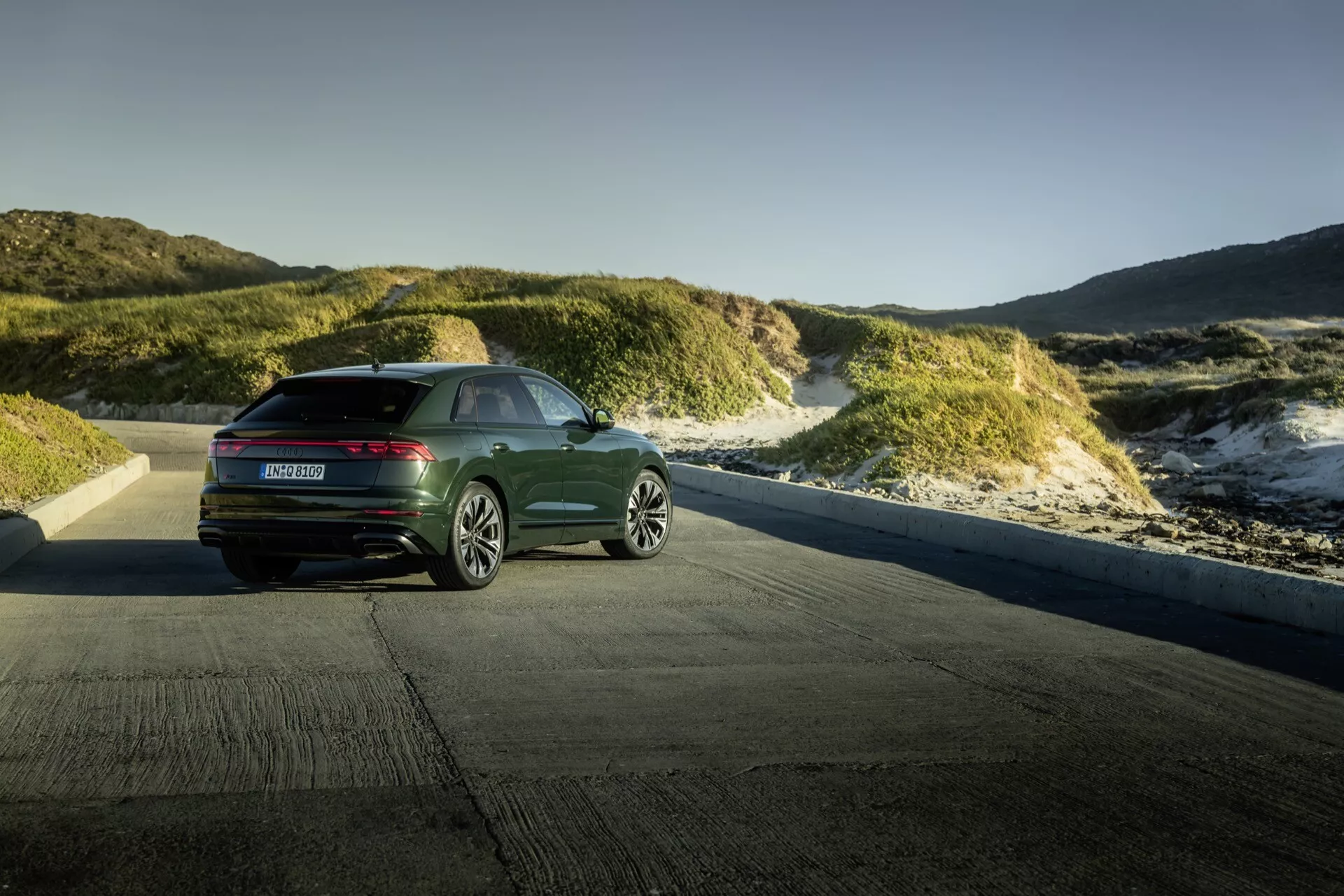 Audi Q8 4M-Facelift 2023 visad mot ett kustnära dynlandskap under gyllene timmen, visar en bakre tre fjärdedelars vy med moderna horisontella bakljus och elegant design