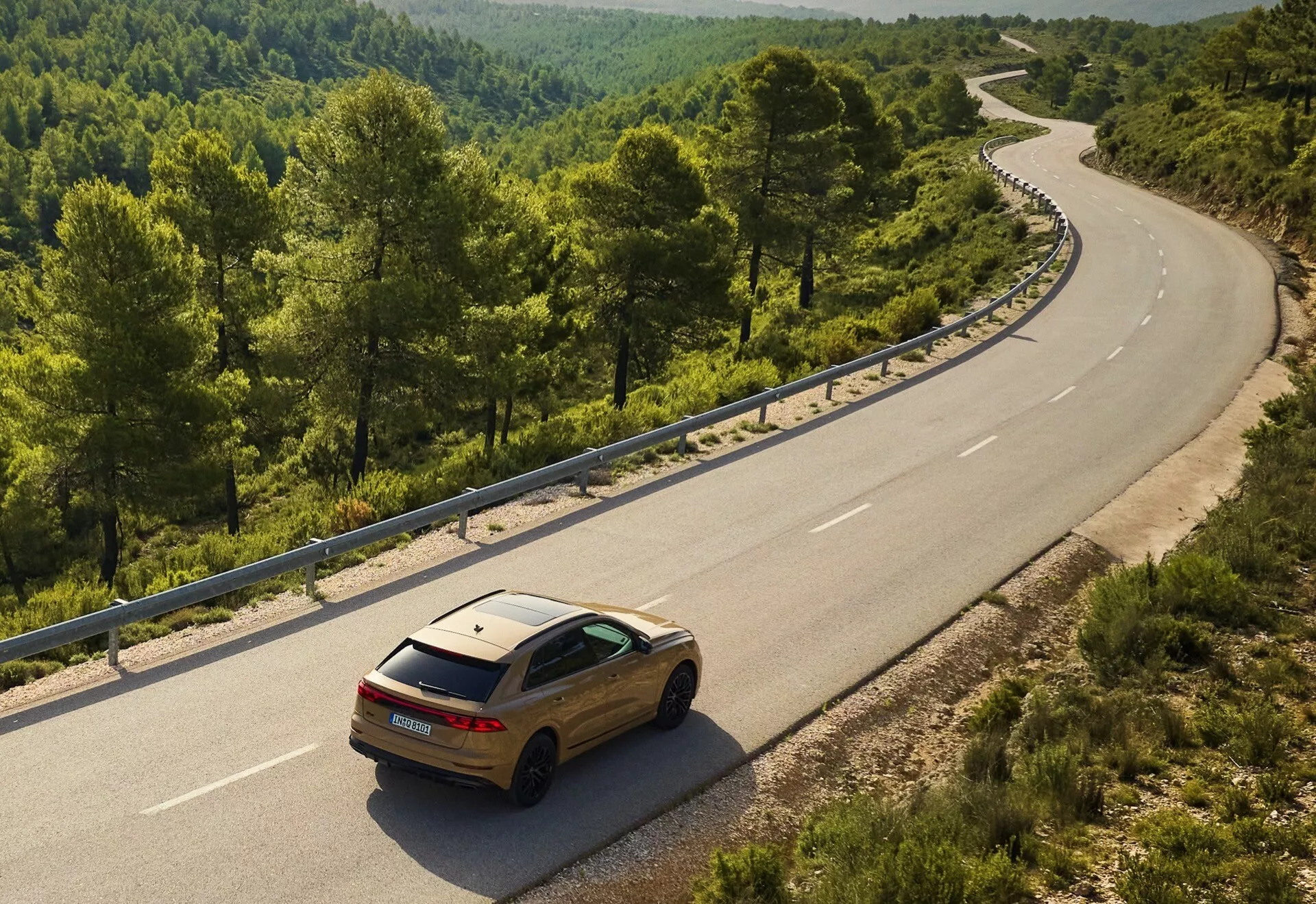 Guldfärgad Audi Q8 2023 (4M-Facelift) på en slingrande tvåfilig väg i en landsbygds- eller skogsmiljö, sedd från en höjd som visar dess bak- och sidodesign mot det naturliga landskapet