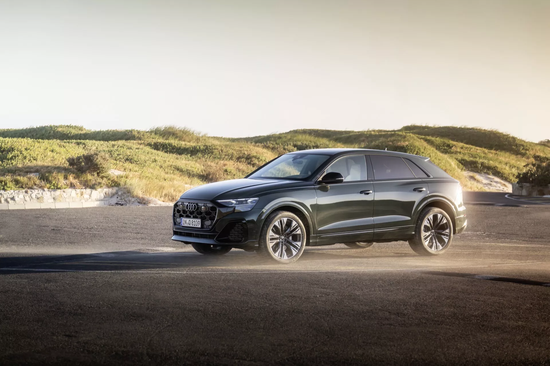 Modern crossover Audi Q8 2023 (4M-Facelift) utomhus under gyllene timmen, som visar avancerade LED-strålkastare, lyxigt grill med säkerhetsfunktioner, prestandainriktade stora lättmetallfälgar, metallisk lack och sportiga designlinjer mot en bakgrund av asfalt och mjuka kullar, vilket framhäver mångsidiga kapaciteter och design