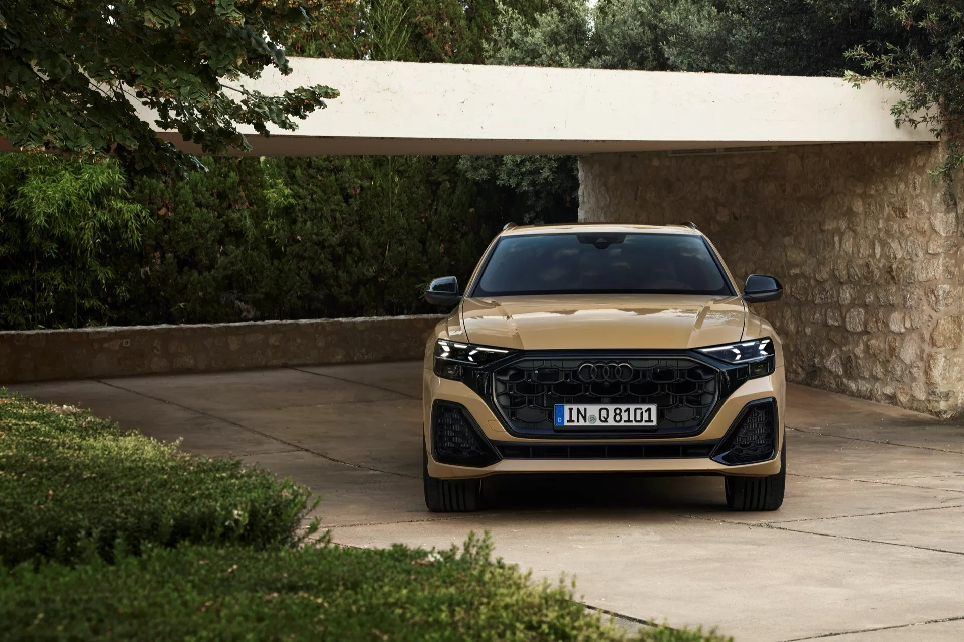Audi Q8 2023 (4M-facelift) visas från en frontal vinkel, med fokus på dess breda grill, slanka LED-strålkastare och metallisk finish, placerad på en uppfart omgiven av frodig grönska