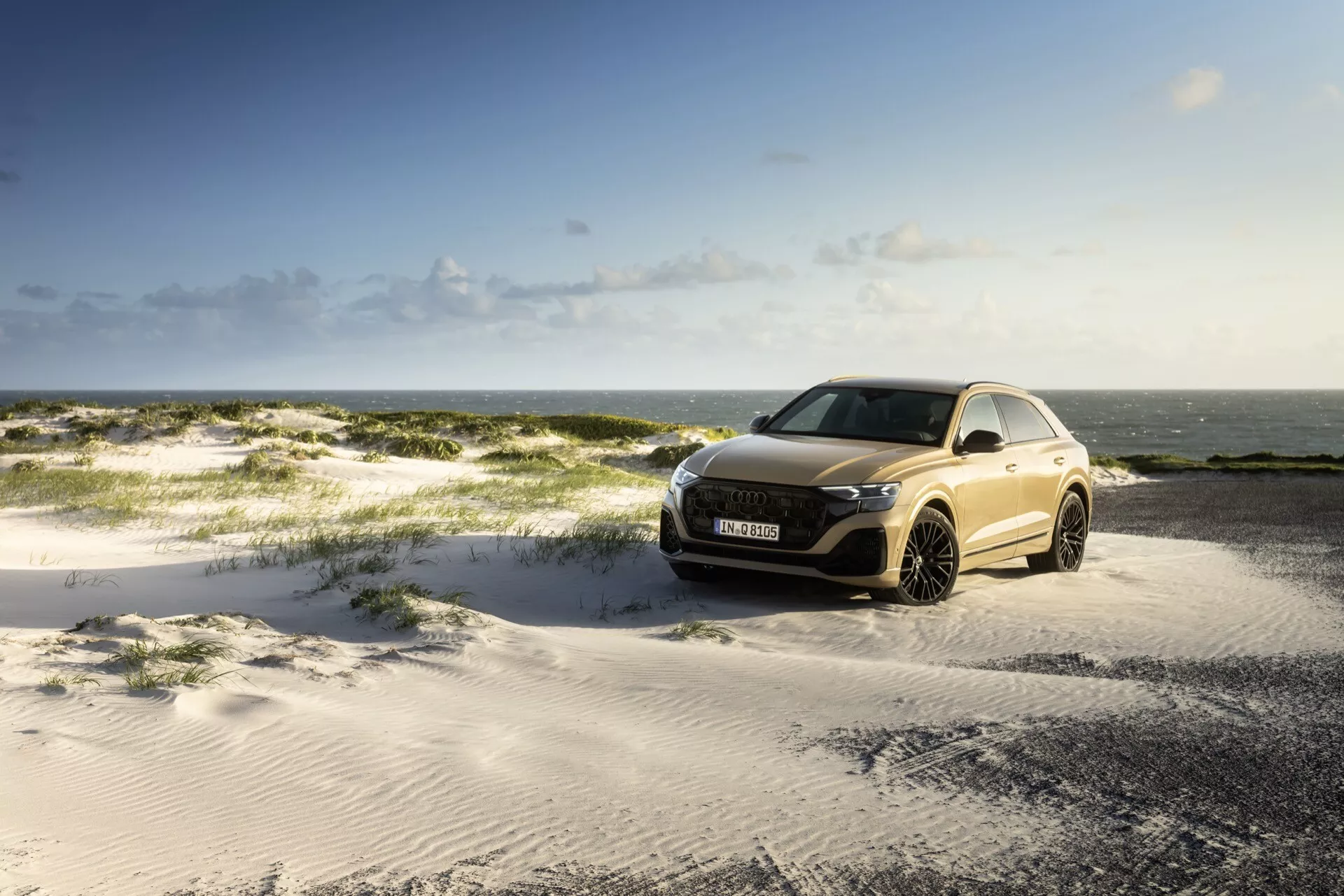 Audi Q8 2023 (4M-Facelift) parkerad på sandig terräng under delvis molnig himmel med havet i bakgrunden, visar främre grillen, LED-strålkastare och aerodynamisk design