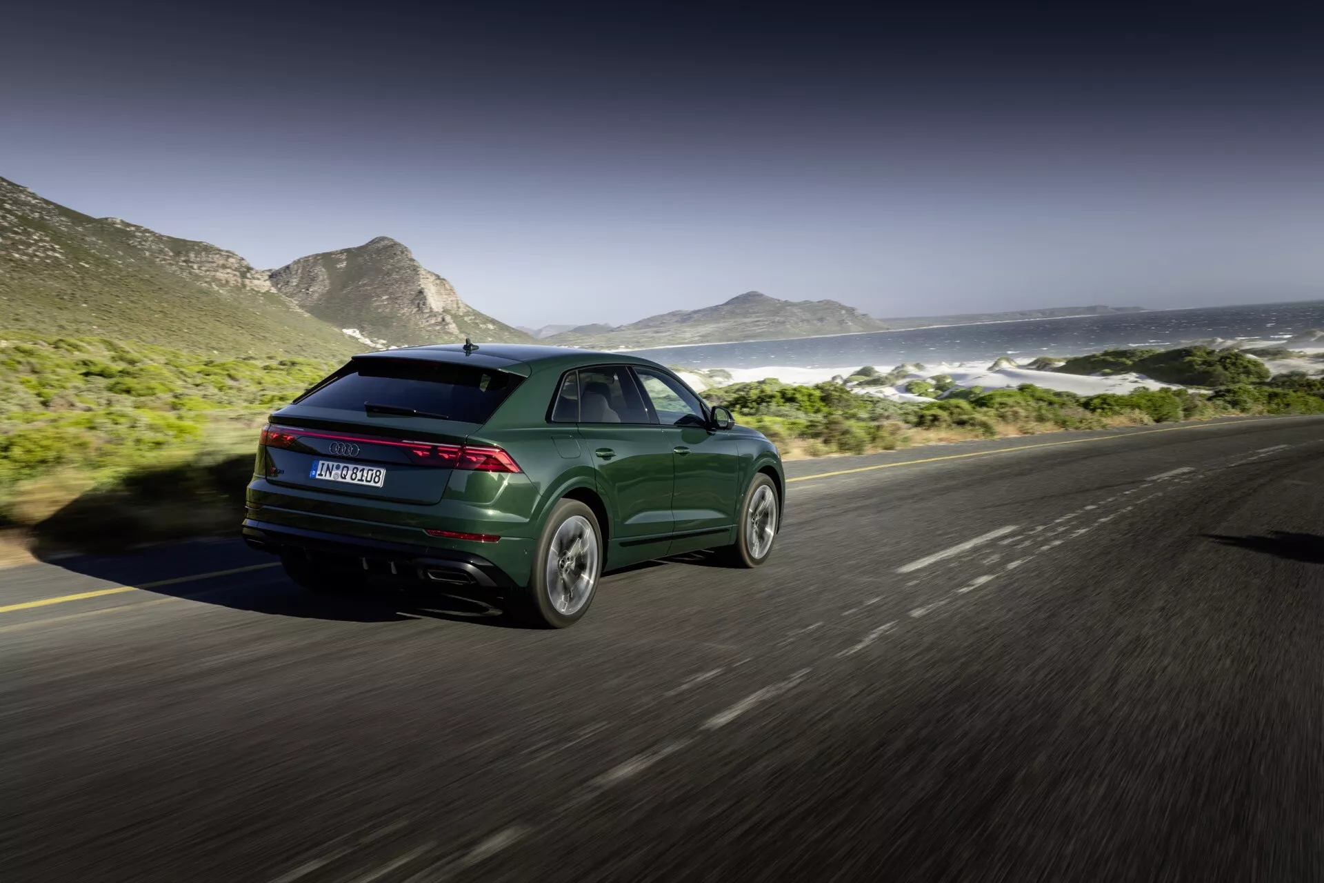 Audi Q8 2023 (4M-Facelift) i metalliskt mörkgrönt, som visar dess sportiga och moderna design med slanka bakljus och lättmetallfälgar, fångat från ett bakre trekvarts perspektiv på en kustväg med berg och hav i bakgrunden
