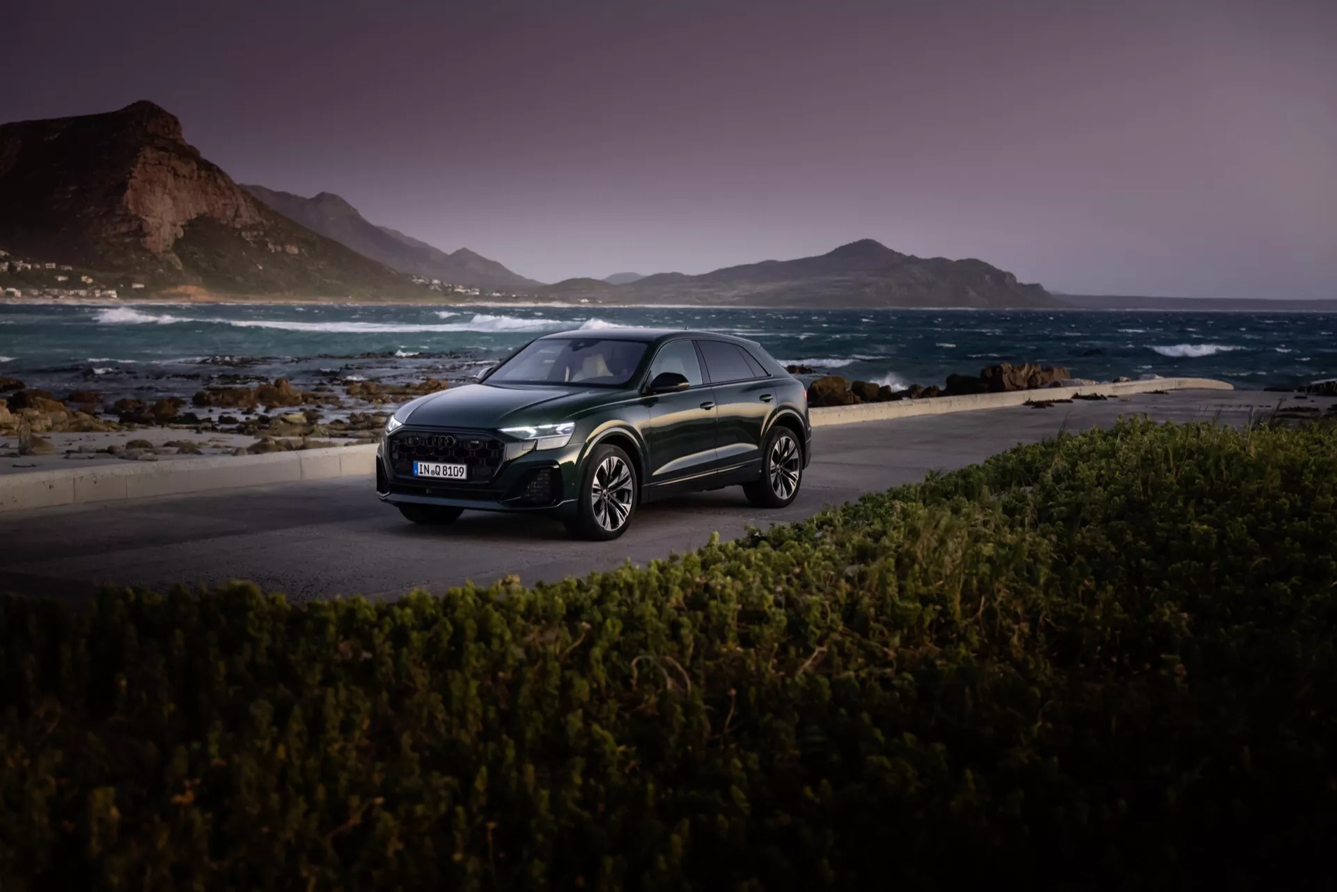 Audi Q8 2023 (4M-Facelift) mörkfärgad halvkombi avbildad på en naturskön kustväg i skymningen, som visar sina LED-strålkastare, aerodynamiska profil och legeringshjul