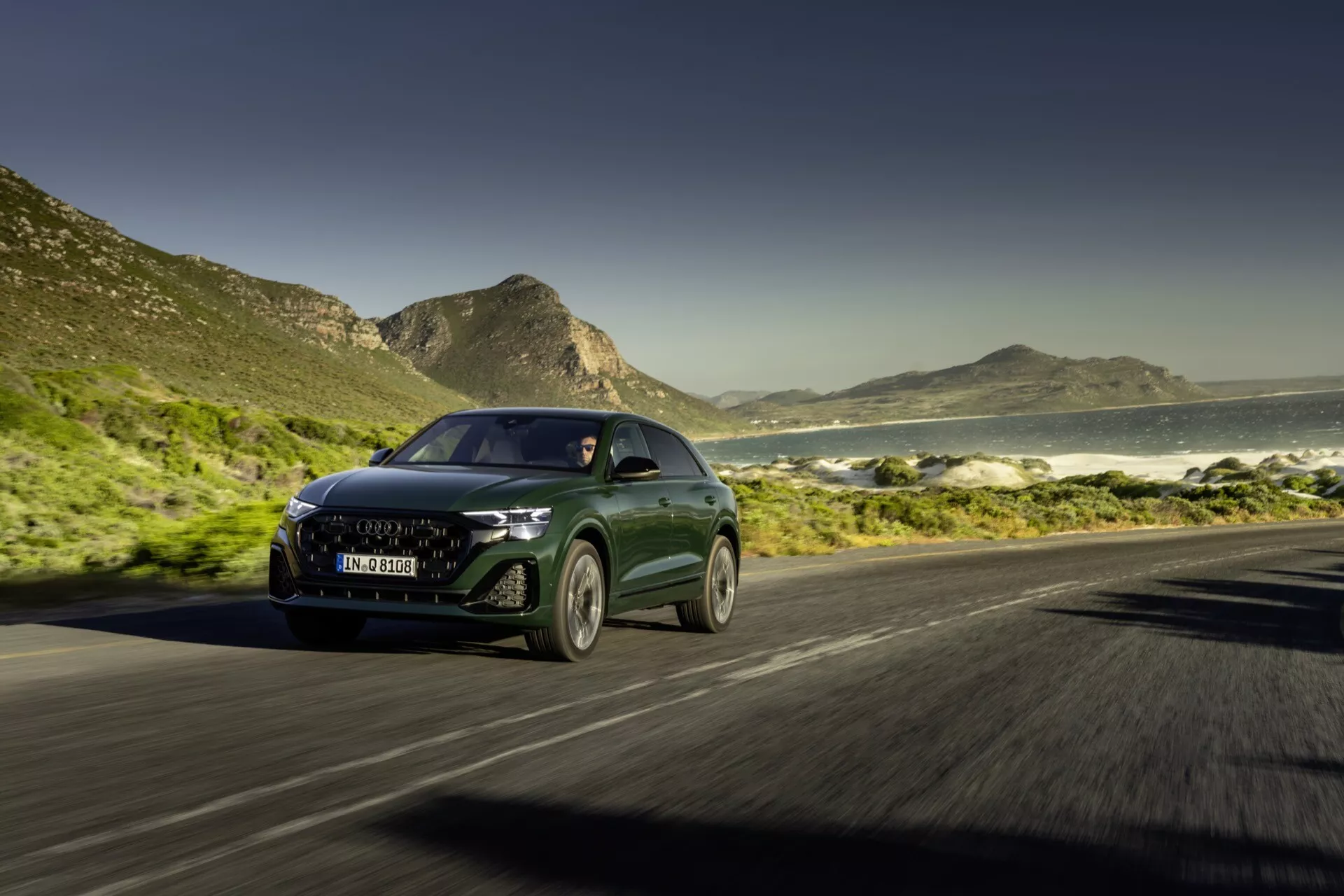 Audi Q8 4M Facelift 2023 kör på kustväg med dynamisk främre tredjedels vy som visar LED-strålkastare, modern grill och rörelseoskärpningseffekt mot en bakgrund av hav och berg under soliga himlar